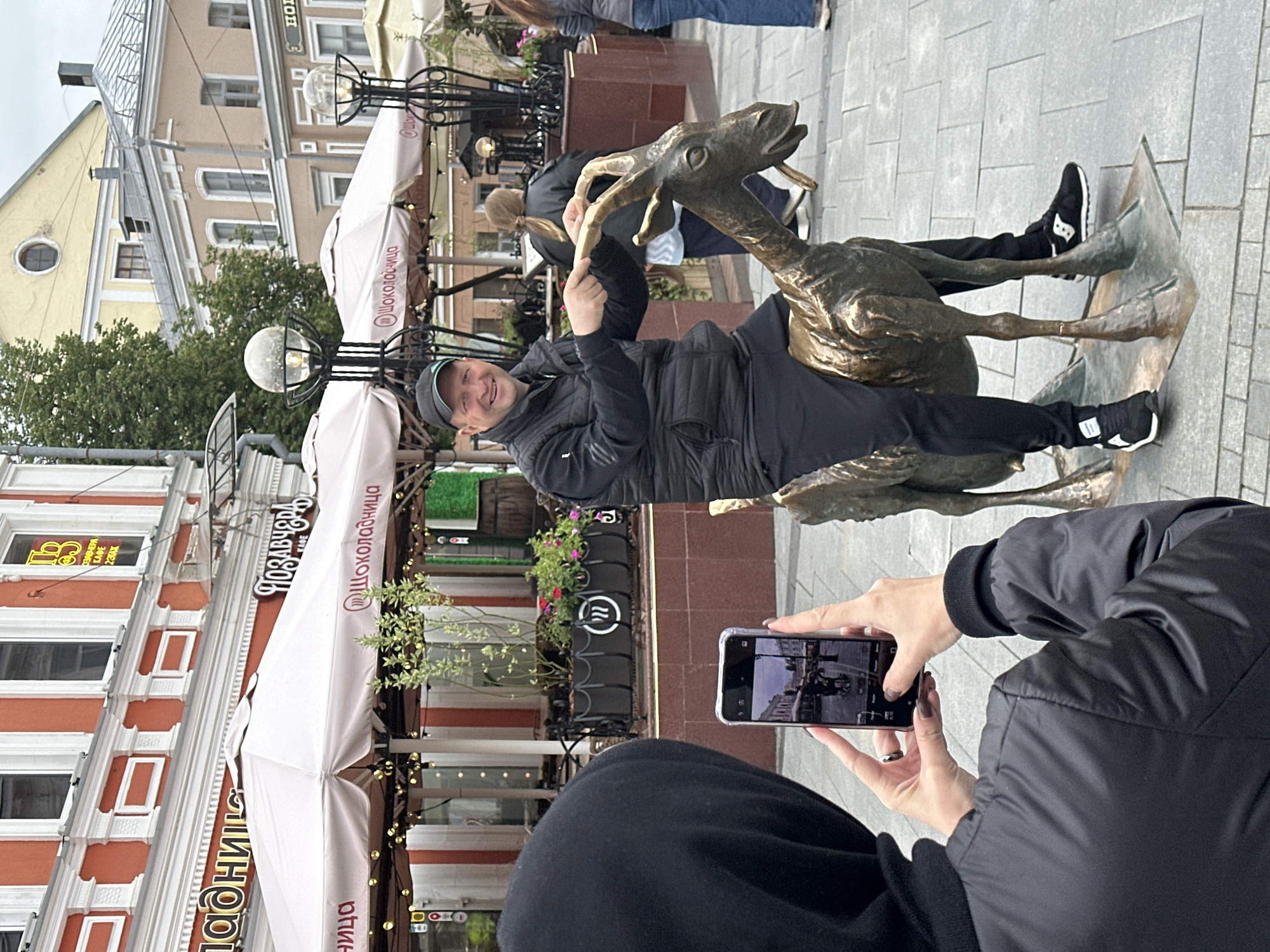 A man is sitting on a bronze sculpture of a deer or goat, smiling and pointing at one of its antlers, while another person takes a picture with their cell phone. The backdrop includes an outdoor cafe or restaurant with light-colored umbrellas. The architecture in the background suggests a European city, with red and white buildings. The cafe appears to be "шоколадница," suggesting this is in Nizhny Novgorod, Russia.