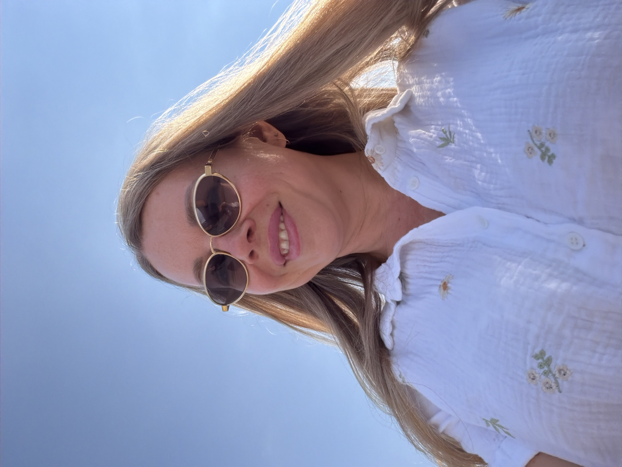 A low-angle, medium shot shows a fair-skinned woman in her late 20s or early 30s, with blonde hair, wearing sunglasses and a white collared shirt with floral embroidery. She is outdoors on a sunny day, with a clear blue sky as the background. Her head is tilted back slightly and she is smiling. Her hair is flowing to her right, catching the sunlight. She is wearing a light-colored shirt with a floral print. The image is a selfie, taken from a slightly downward angle. The location is stated to be Reggello, Italy.