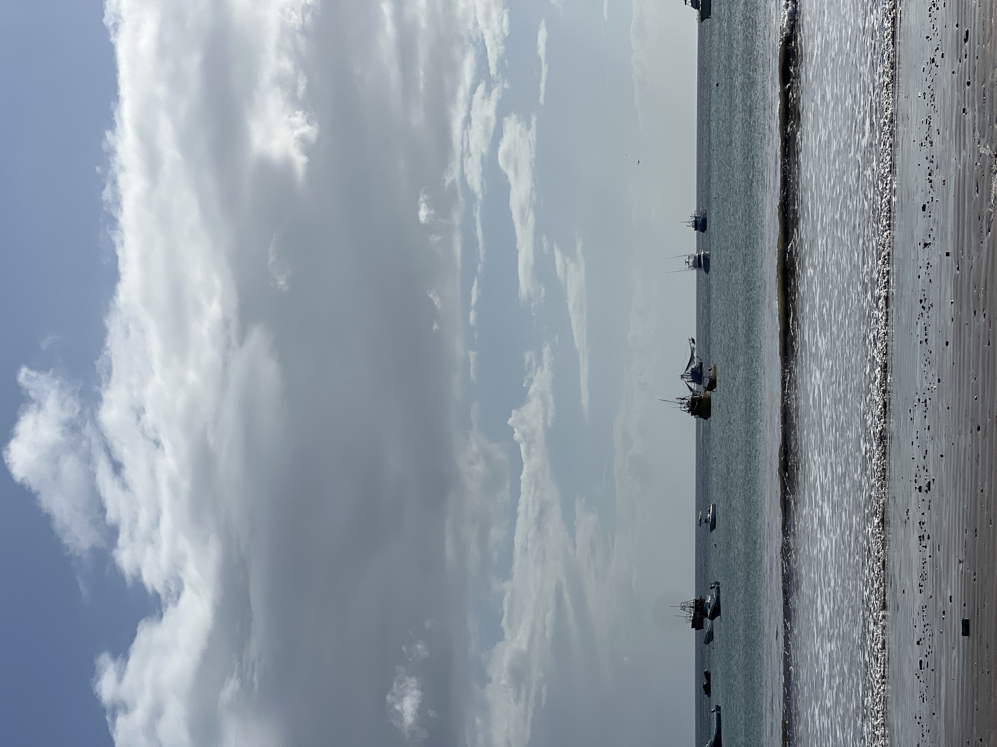 A tranquil scene unfolds in San Juan del Sur, Nicaragua, showcasing a beach with calm waters and a fleet of boats at anchor. The sand, a mixture of dark grains and small pebbles, stretches towards the water's edge, where gentle waves create a foamy white line. Further out, the sea is a muted blue-green, dotted with numerous fishing boats of various sizes. These vessels, some with tall masts and rigging, are positioned at different distances from the shore, suggesting a working harbor. Above, the sky is a canvas of dramatic clouds, with patches of bright blue peeking through. Some clouds are white and wispy, while others are darker and more imposing, hinting at changeable weather. The overall atmosphere is one of peaceful coexistence between human activity and the natural elements of the coast.