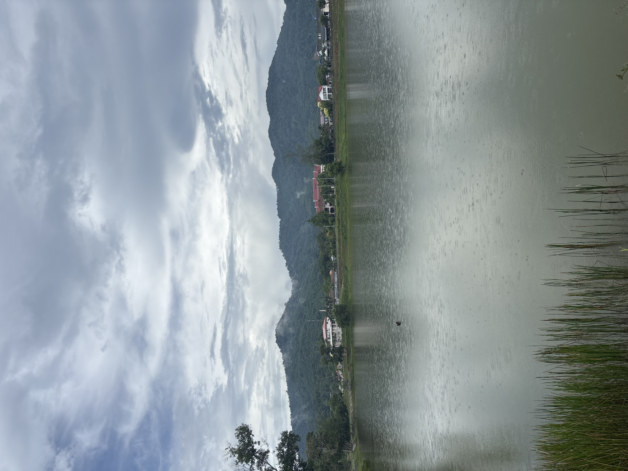 The image depicts a rainy day at a lake in Choeng Thale, Thailand. A mountain range borders the water's edge, dotted with scattered houses and trees. The sky is overcast with dense, striated clouds. Rain falls on the lake surface, creating a textured, grey appearance. Reeds are visible along the immediate shoreline. A single, dark object floats in the water.