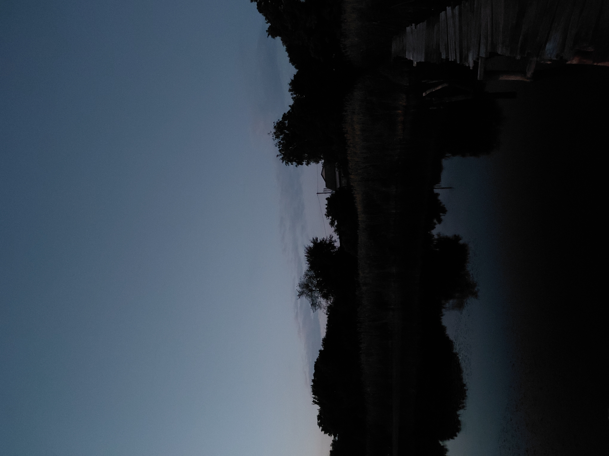 The image depicts a twilight scene of a calm body of water with a wooden pier extending from the right side into the water. The sky is a clear, deep blue, transitioning to a lighter shade towards the horizon, with faint wisps of clouds visible. A dense line of silhouetted trees and vegetation forms the backdrop across the horizon, with a small house visible amongst the trees on the left side. The water reflects the dark silhouettes of the trees and the sky, creating a dark, almost black surface with subtle ripples. The wooden pier appears weathered and is constructed from rough-hewn planks. The overall atmosphere is serene and still, characteristic of dusk. The location is identified as Ustyvytsia, Ukraine.