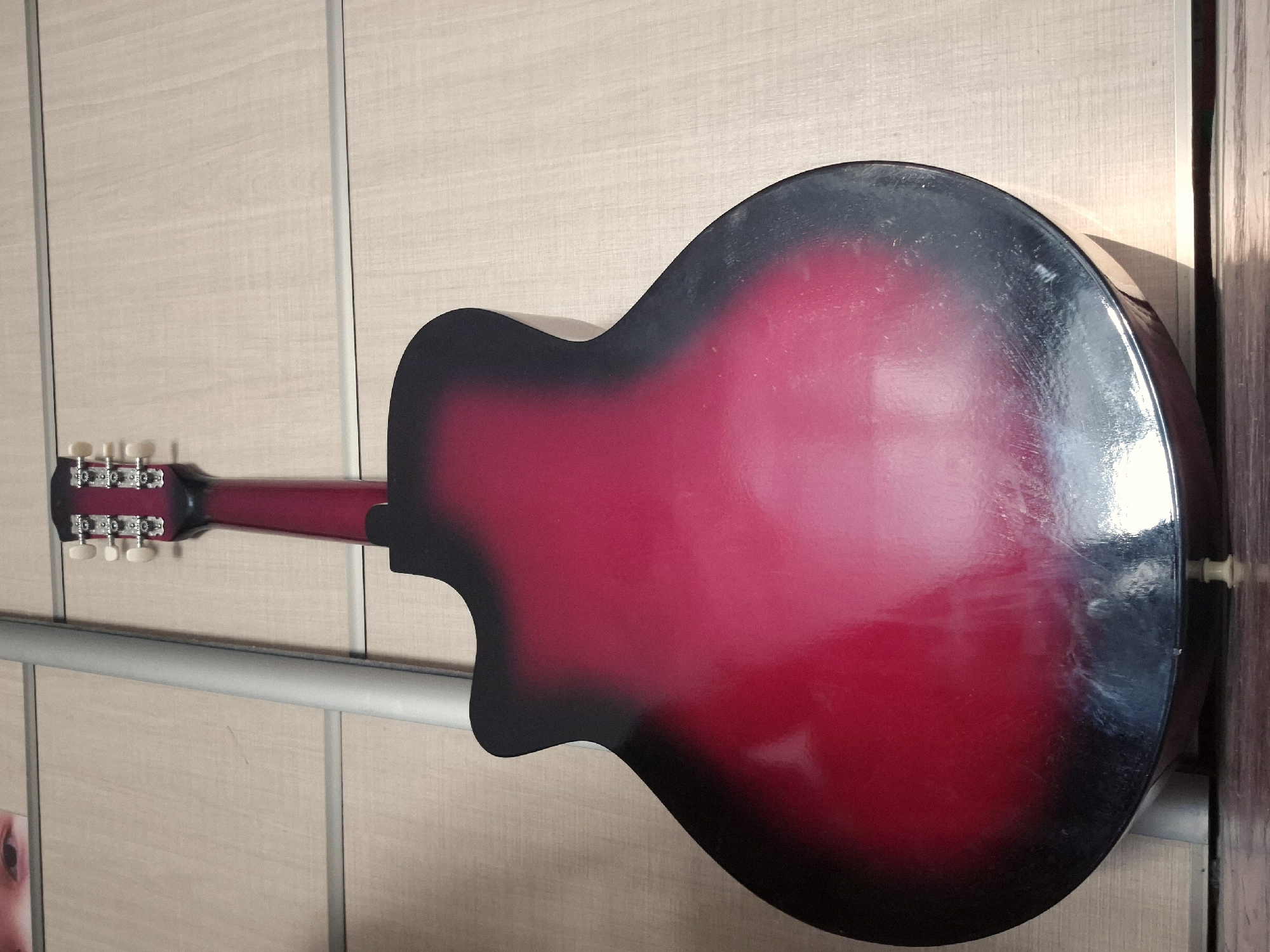 A red and black acoustic guitar is leaning against a light-colored wall with vertical paneling. The guitar is positioned vertically, with its headstock at the top and its body at the bottom. The headstock has six tuning pegs. The body of the guitar is a gradient of bright red fading to black at the edges. A silver pole runs vertically alongside the left side of the guitar. In the upper left corner of the image, a portion of a person's face is visible, with one eye looking towards the camera. The scene appears to be indoors, possibly in a room or closet, given the wall paneling and the presence of a metallic pole which could be part of a closet rod. The lighting suggests an indoor environment with some natural or artificial light source. No specific location in Kokshetau, Kazakhstan can be determined from the image.