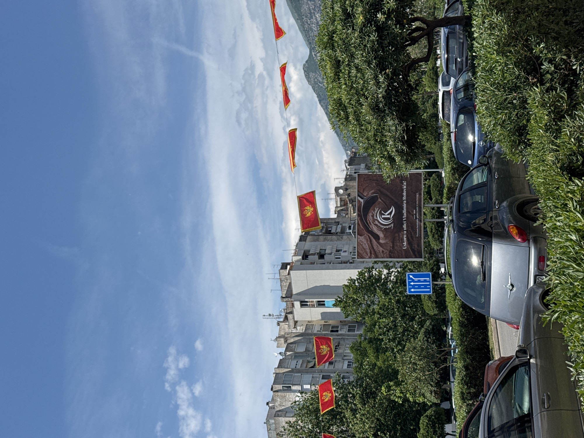 The image depicts a street scene in Bjeliši, Montenegro. Several Montenegrin flags are strung across the street. A building with a large advertisement for "Uskoro u ulici Vladimira Rolovica!" is visible. Cars are parked along the side of the street. The sky is blue with scattered clouds, and a mountain range is in the background. Trees and bushes line the street. A blue road sign is also visible.