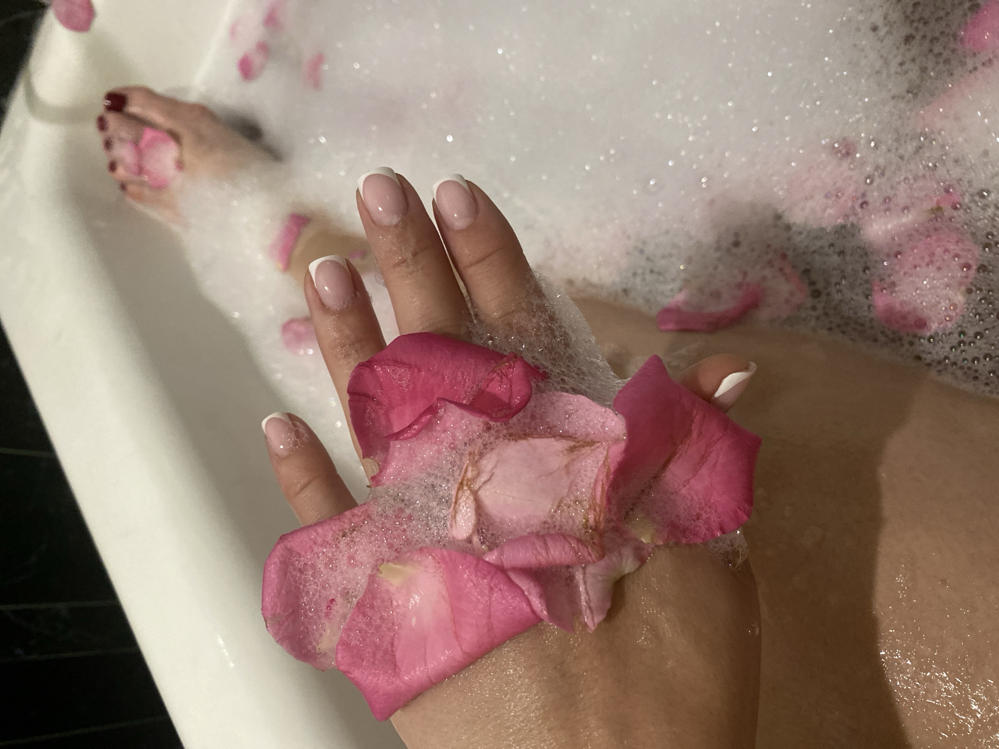 The scene depicts a person in a bubble bath filled with rose petals. The individual's hand is prominently displayed, holding several pink rose petals covered in bubbles. The hand exhibits a fresh manicure with white tips. A foot with red nail polish is also visible in the upper left corner of the frame. The background is filled with white bubbles and scattered pink rose petals. The location in Sochi, Russia is not evident from the visual content of the image.
