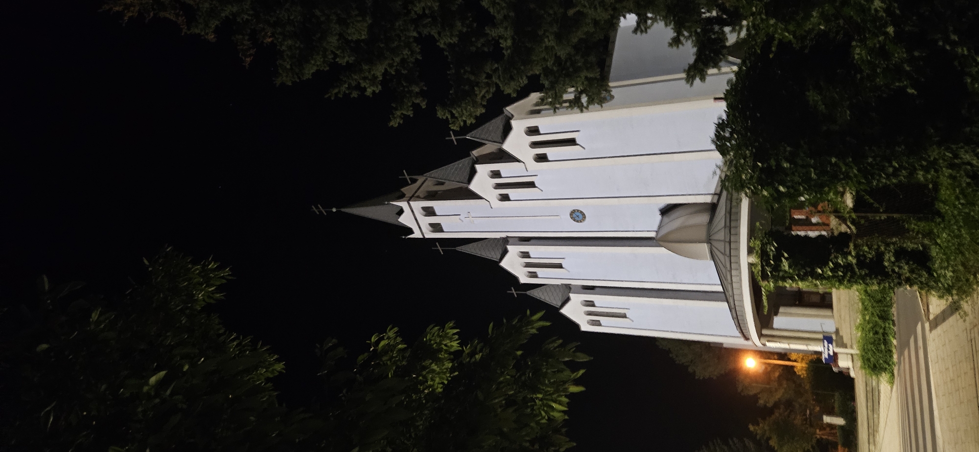 An exterior shot captures a church at night in Hévíz, Hungary. The church is lit by artificial lights, highlighting its pale blue facade and multiple pointed spires topped with crosses. A clock face is visible on the central tower. The foreground is partially obscured by dark green foliage, with trees framing the scene on the sides. A lamppost with an orange glow is visible to the left of the church, and a paved walkway leads towards the entrance. The sky above is dark and clear, suggesting nighttime.