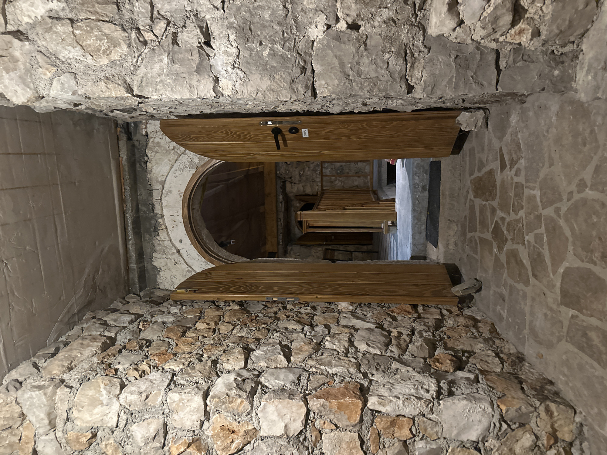 The image shows a stone corridor in Pečurice, Montenegro, with rough-hewn stone walls and a stone-paved floor. An arched doorway with wooden doors stands open, leading into a dimly lit interior. The doors are made of light-colored wood and have black hardware. One door has a small white sign with black lettering that reads "GURAU". Beyond the doorway, a wooden railing and stairs are visible. The ceiling appears to be concrete. The scene is devoid of people.