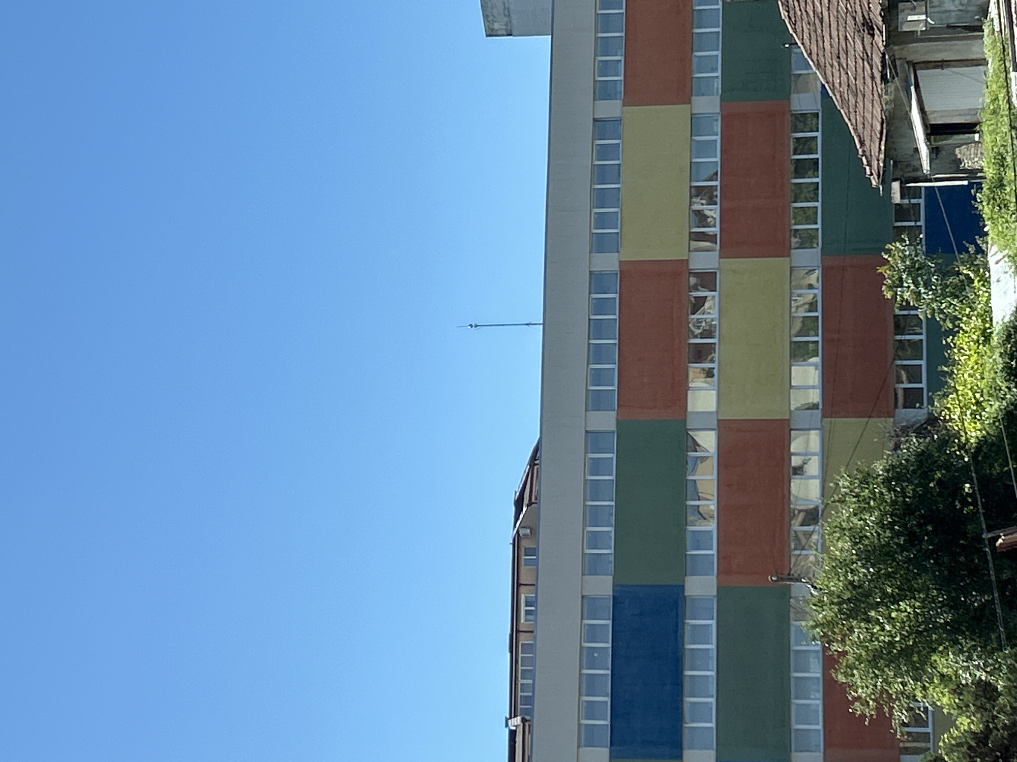 The image shows a tall, colorfully-painted building in Sofia, Bulgaria, situated under a clear blue sky. The building is divided into vertical sections of yellow, red, green, and blue panels separated by continuous white lines. A slender antenna extends from the top of the building. Trees are visible on the right side of the building.