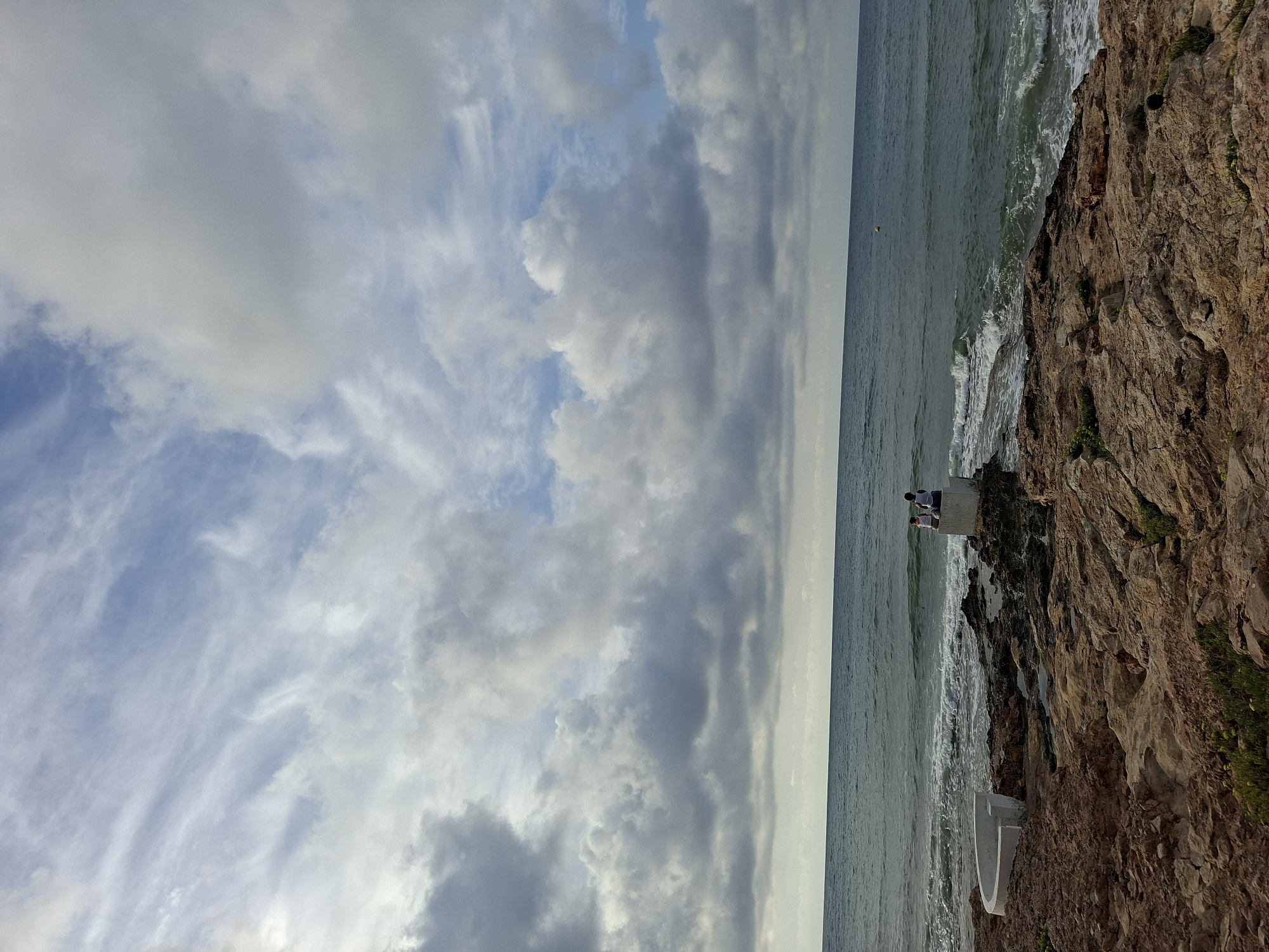 The image depicts a rocky coastline along the Mediterranean Sea in Torrevieja, Spain. The sky is filled with a mix of wispy and cumulus clouds, with patches of blue sky visible. The sea is a deep blue-green, with moderate waves breaking on the shore. In the foreground, the coastline is characterized by reddish-brown rocks with sparse vegetation. A white, curved structure, possibly part of a coastal defense or seating area, is visible on the left. Further along the coast, two individuals are seated on a square concrete block, facing the sea. One person is wearing a white tank top and the other a dark shirt. The overall scene suggests a quiet, natural coastal landscape.