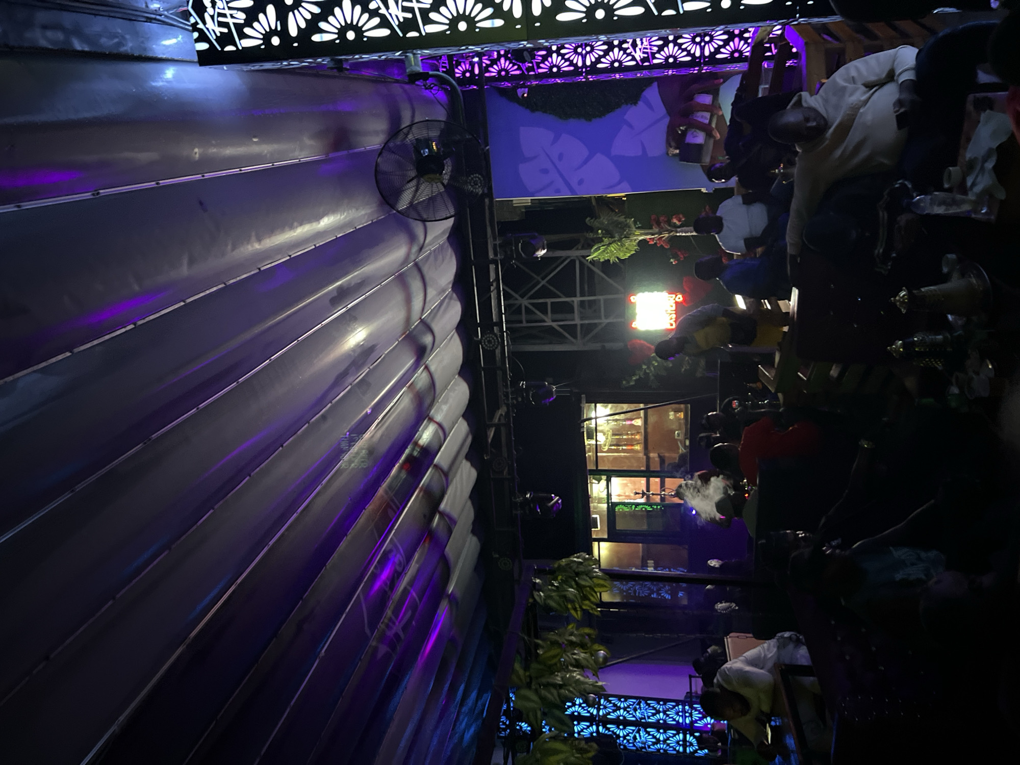 The image depicts an outdoor seating area of a hookah lounge, likely in Howayi, Nigeria, illuminated by purple and blue lighting. Several individuals are seated on couches and chairs, some engaging with their phones, while others appear to be conversing. Hookah pipes are visible on tables, with smoke emanating from one. The environment is decorated with hanging plants and patterned screens. A ceiling fan is mounted above the seating area. The overall atmosphere suggests a relaxed, social gathering.
