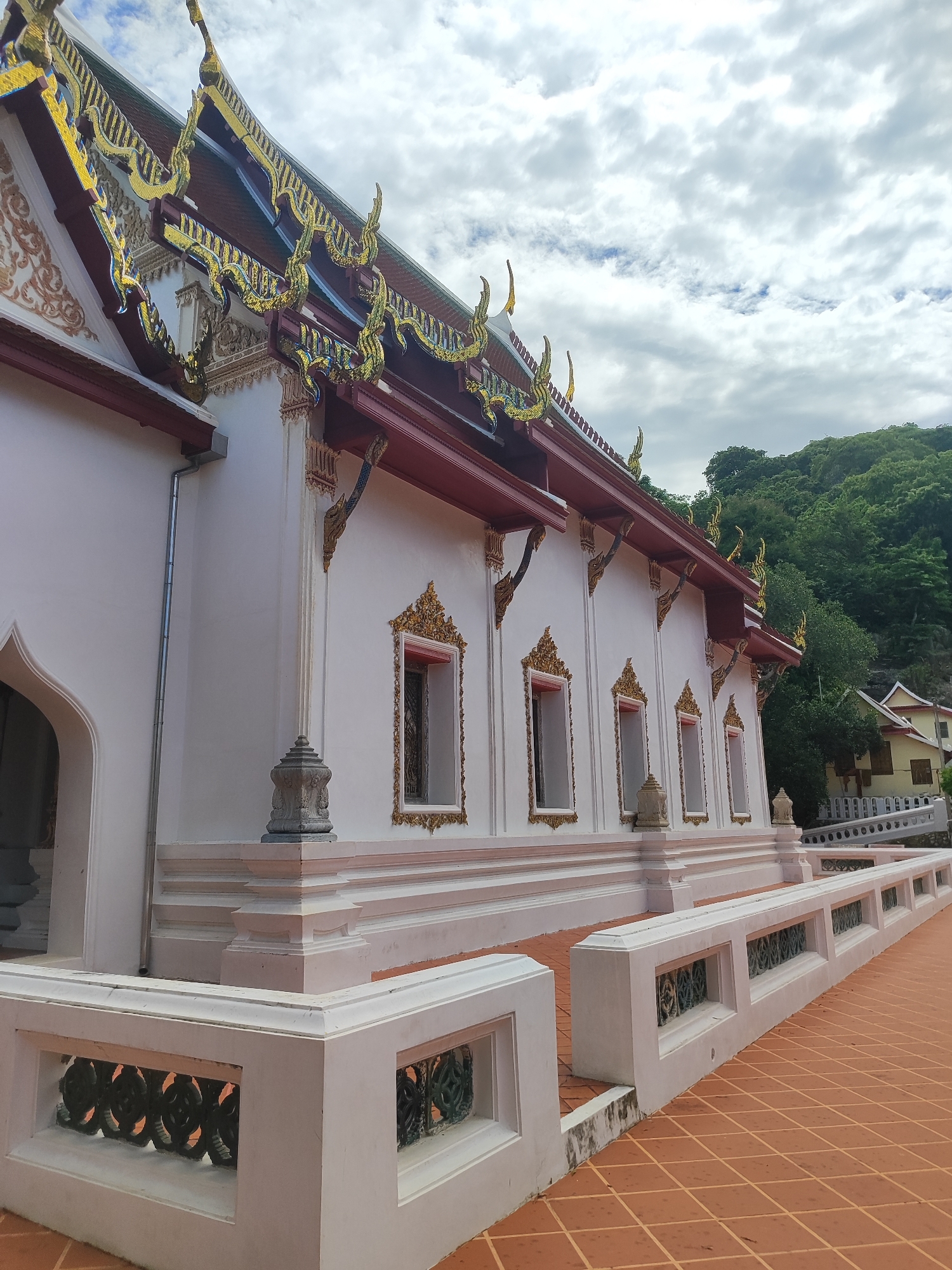 This image shows the exterior of a Buddhist temple, likely a Wat, in Phetchaburi, Thailand. The temple features traditional architecture with a white facade, intricate gold and red roof detailing, and decorative window frames. A tiled walkway leads away from the temple, bordered by a white balustrade with decorative metalwork. Lush green hills form the backdrop, and the sky is filled with dramatic clouds. The scene is captured from a low angle, emphasizing the scale and ornate design of the temple structure. No people are visible in the image.