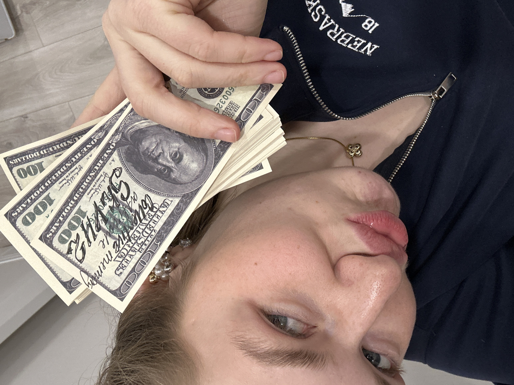 A woman is holding a stack of what appear to be US $100 bills near her head. Some of the bills have writing on them. She is wearing a navy blue zip-up jacket with "NEBRASKA 18" embroidered on the chest, along with a gold necklace. The setting appears to be indoors. There is no evidence to suggest this photo was taken in Kazan, Russia.