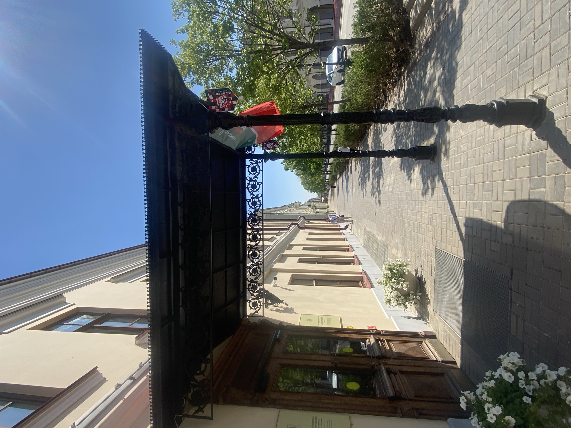 This image shows a street in Kazan, Russia. The camera is positioned near the entrance of a building with a dark metal awning decorated with intricate patterns. The door is made of wood, with glass panels reflecting the surroundings. A sign is visible near the door, but its details are unclear. Flower pots with white flowers are placed along the building's exterior. A row of black lampposts lines the walkway. Flags and banners, including an "AK BARS" emblem, are attached to the lampposts. The street is paved with rectangular blocks. Trees are visible in the background. The sky is clear and blue. There are no people visible.