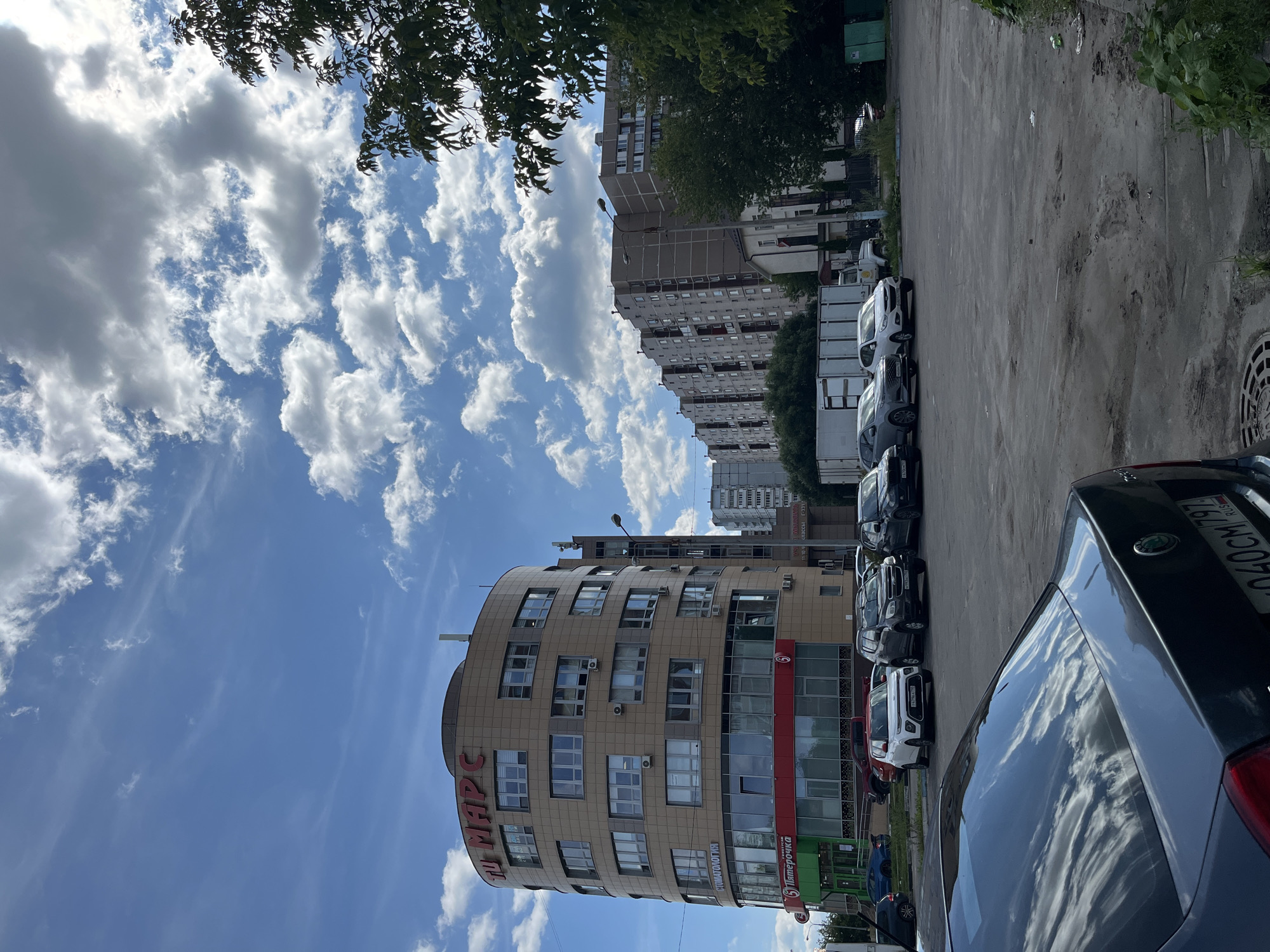 This is a daytime, eye-level, outdoor, wide shot of a city street scene in Balashikha, Russia. The sky is blue with scattered white and gray clouds. In the foreground, the rear of a dark-colored car is visible, with reflections of the clouds on its window. To the left and center of the image is a modern, multi-story, rounded beige building with numerous windows and air conditioning units. The building has red signage at the top that reads "Ю МАРС" and a red band on the lower floors with green signage of a grocery store labeled "Пятёрочка". To the right of this building, there is a row of apartment buildings with gray facades and many windows. Trees and foliage are visible on the right side of the frame, and a white truck and several cars are parked in a lot between the buildings and the foreground car. The ground in the foreground and mid-ground is a gray, paved surface, with some areas appearing worn and sandy.