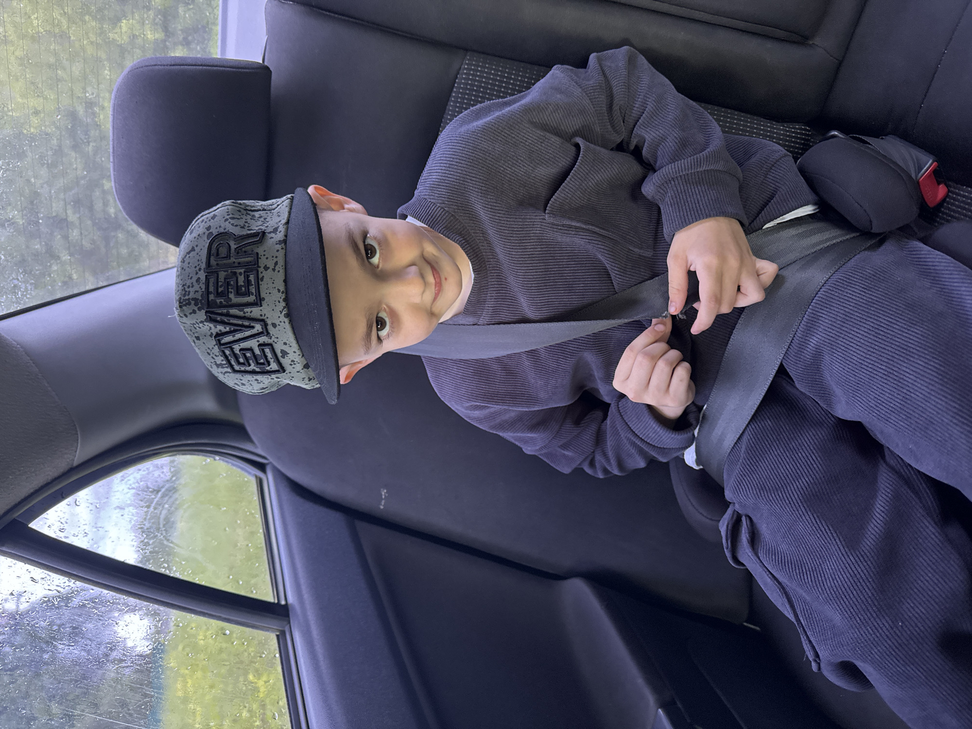 A young child wearing a dark gray corduroy outfit and a gray and black baseball cap with "EVER" printed on it is sitting in the back seat of a car. The child is smiling and appears to be fastening their seatbelt. The interior of the car is dark gray and black. Rain droplets are visible on the car window. It is not possible to ascertain if the image was taken in Shevlyakovo, Russia.