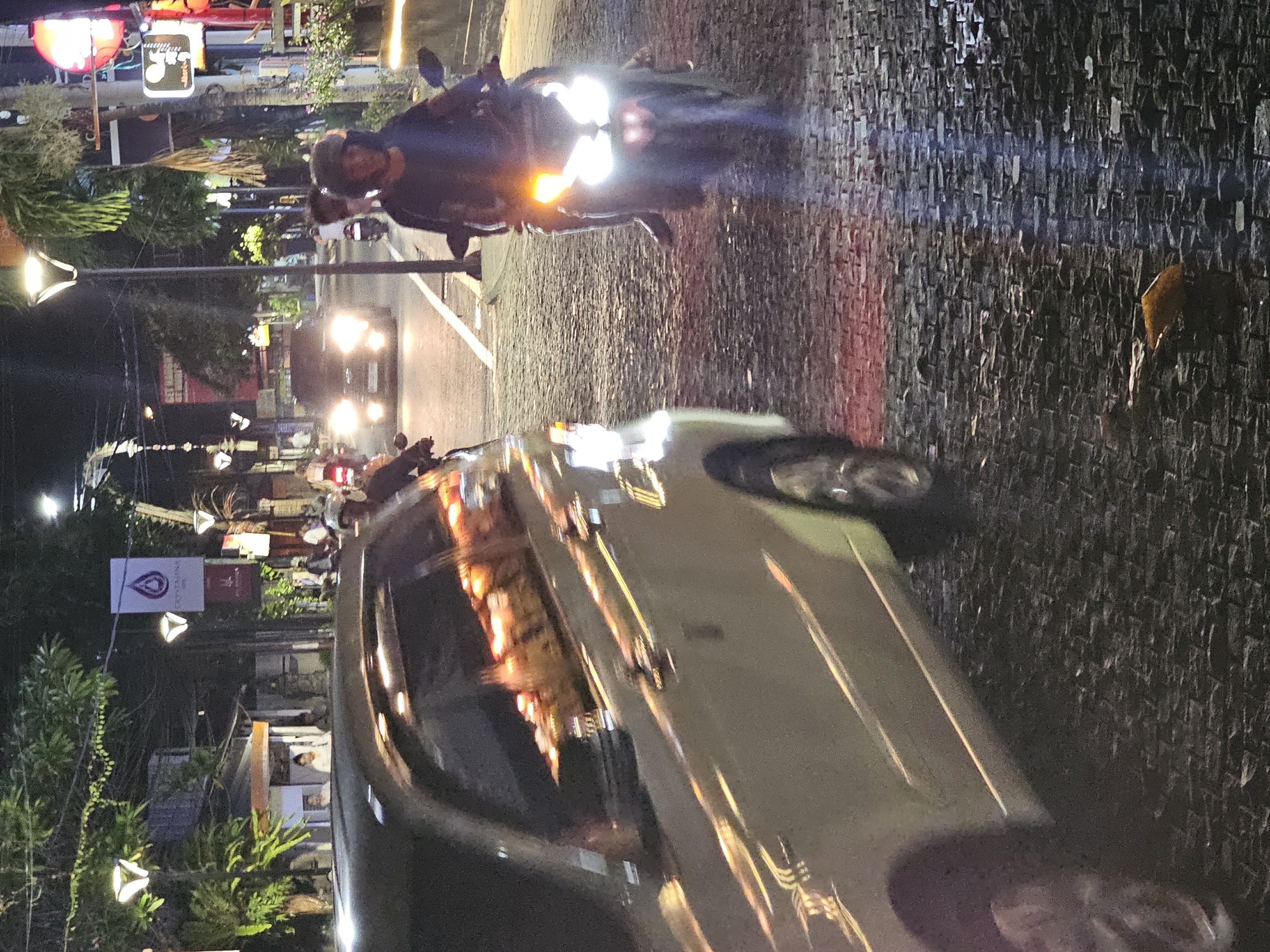 The image, taken at night in Denpasar, Indonesia, shows a street scene with a car and a motorcycle in the foreground. The car, light-colored, appears to be moving slowly, with its headlights reflecting off its surface. A motorcycle, with its headlights on, is positioned behind and to the side of the car, also apparently in motion. The street is paved with cobblestones. Background details include buildings, trees, streetlights, and a sign with a logo in white. Further back, more vehicles are visible with their headlights on. The setting is urban, likely a street in a busy area of Denpasar.