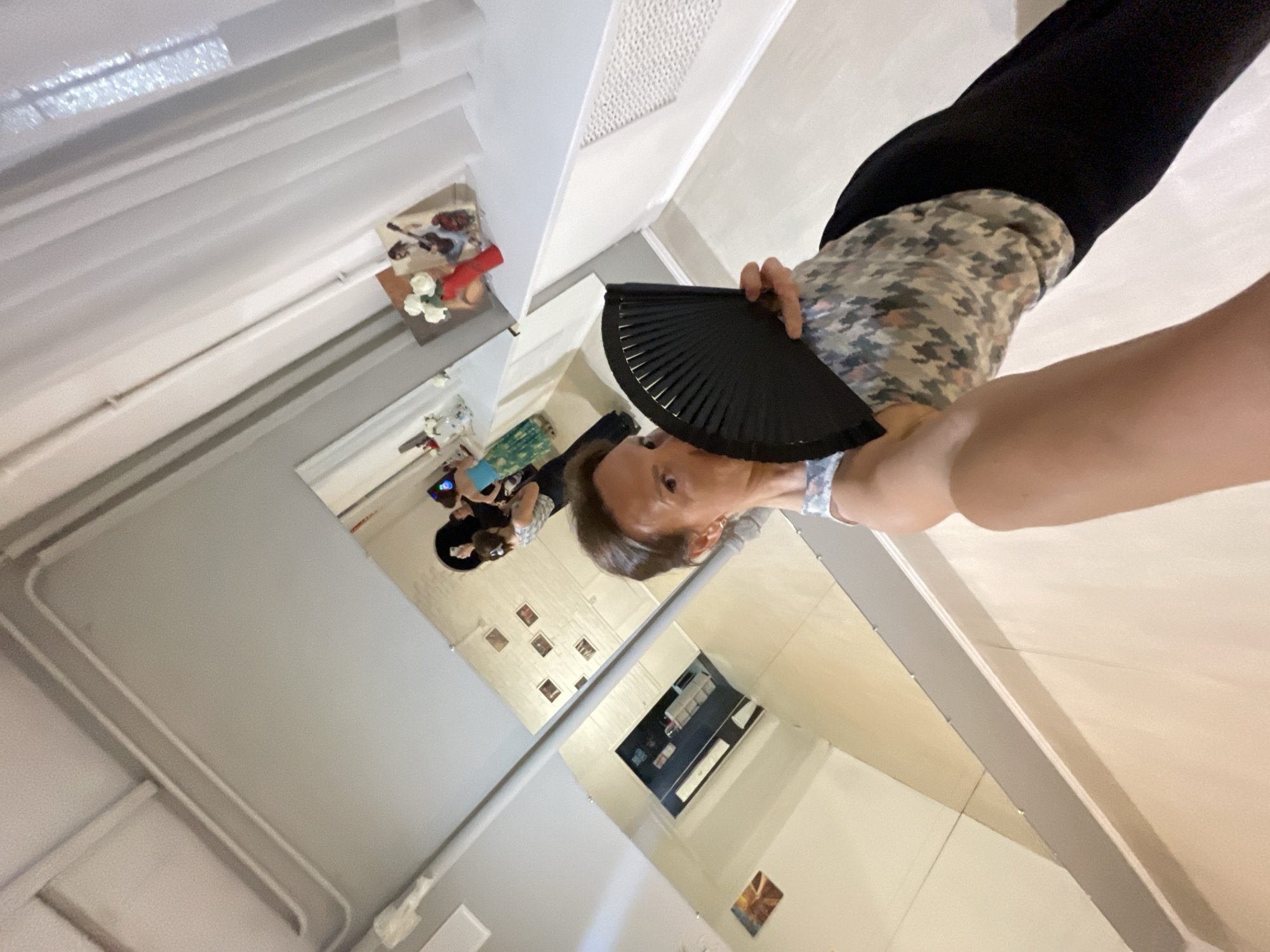 The image shows a woman holding a black fan in the foreground, obscuring part of her face. She is wearing a patterned scarf and black pants. In the background, reflected in a large mirror, are several other people, including a woman in a blue dress and another woman in a striped top. There are also some decorative items, such as vases of flowers and framed pictures, visible in the reflection. The environment appears to be an indoor space with light-colored walls and floors, possibly a dance studio or a similar public venue. There is no indication of the specific location in the city of Sochi, Russia.