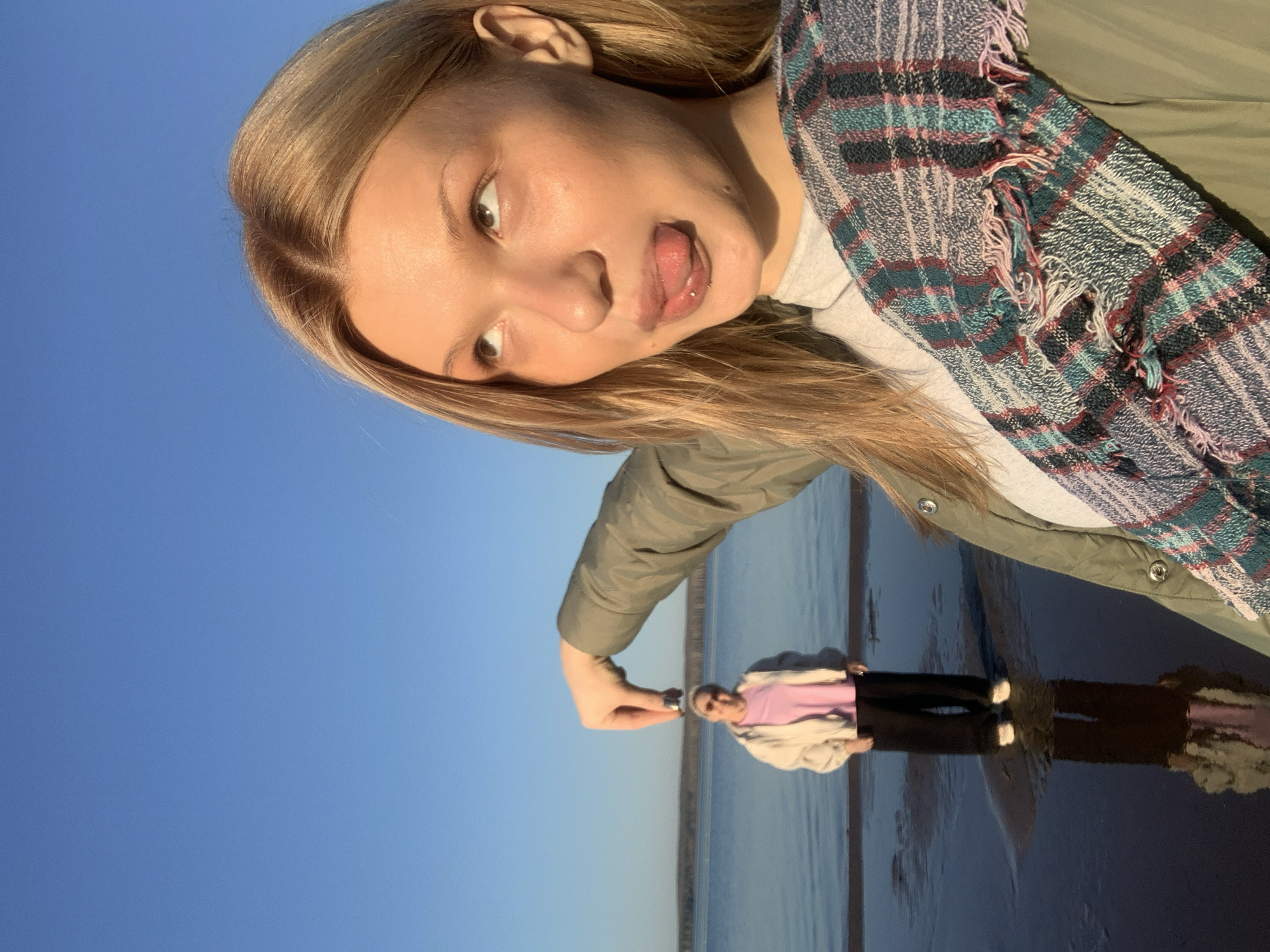 The image shows a young woman posing for a selfie on a beach in Chaikovsky, Russia. She is blonde with fair skin and is sticking out her tongue. She is wearing a green jacket and a plaid scarf. In the background, there is water and a reflection of a woman on the water. The sky is blue and clear. The image appears to be taken near the Kama River, a prominent landmark in Chaikovsky. The action involves the woman taking a selfie and interacting with the environment by posing near the water. The composition includes the woman, her reflection, the water, the sky, and the horizon line.