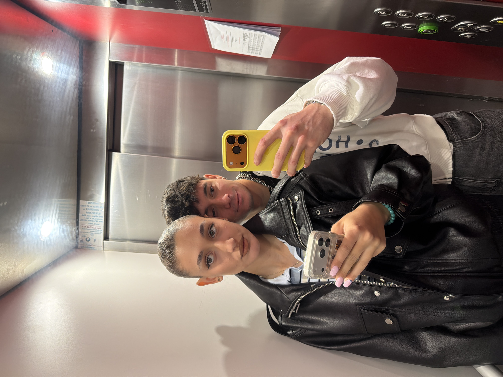 Two people are taking a selfie in what appears to be an elevator. The woman in the foreground wears a black leather jacket and holds a silver iPhone. Her fingernails are painted light pink. A man is visible behind her, holding a yellow iPhone. He's wearing a white sweatshirt and black jeans. The elevator's interior is stainless steel with red accents at the top. A panel of elevator buttons is visible at the top of the frame. The location is not discernible based on the provided image. No definitive evidence points to Sofia, Bulgaria.