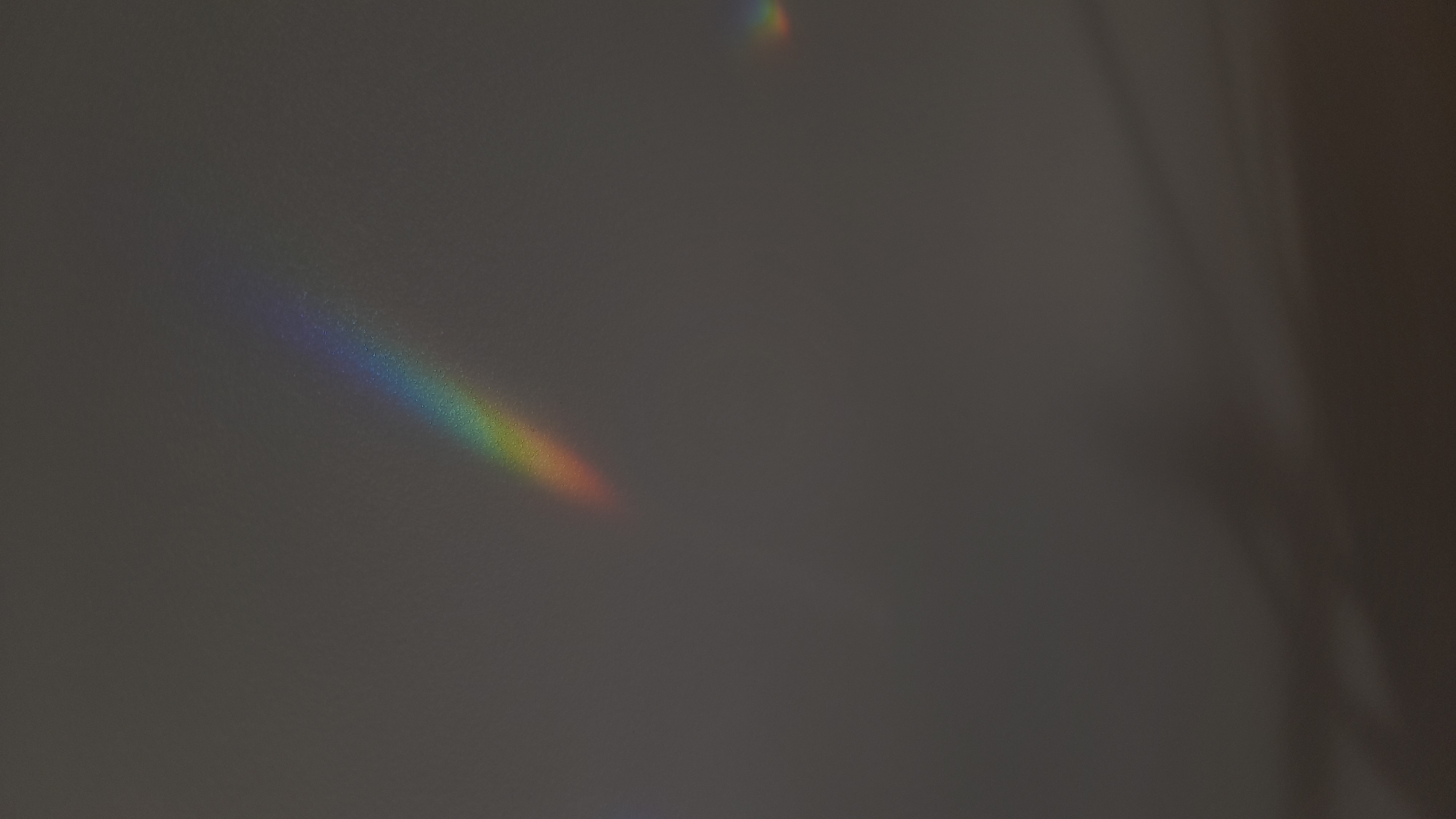 The image depicts a gray, textured surface, possibly a wall, with a faint rainbow spectrum projected onto it in the lower left quadrant. A smaller, fainter rainbow appears near the top center. To the right, the surface transitions to a darker shade with subtle tonal variations, suggesting a rounded edge or corner. The setting appears to be an interior space, and there's no indication of Saint Petersburg, Russia.