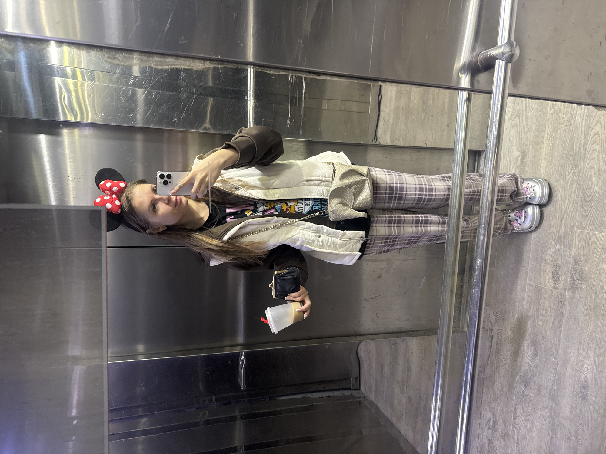 A female individual is captured in a mirror selfie within an elevator. She stands centrally, holding a silver-colored smartphone in her right hand to take the photograph. On her head, she wears a black headband with Minnie Mouse ears and a red polka-dotted bow. Her attire includes a black graphic t-shirt, a brown long-sleeved layer, and a cream-colored quilted sleeveless vest. She wears light plaid trousers and silver clogs with pink decorative charms. In her left hand, she holds a clear plastic cup containing a light brown beverage and a small black coin purse. A light-colored, elephant-shaped crossbody bag with a chain strap is worn over her right shoulder. The elevator interior consists of brushed metal walls displaying numerous scratches and reflections, and a light-colored floor with a wood-grain pattern. A double-barreled metal handrail extends horizontally across the lower section of the scene.