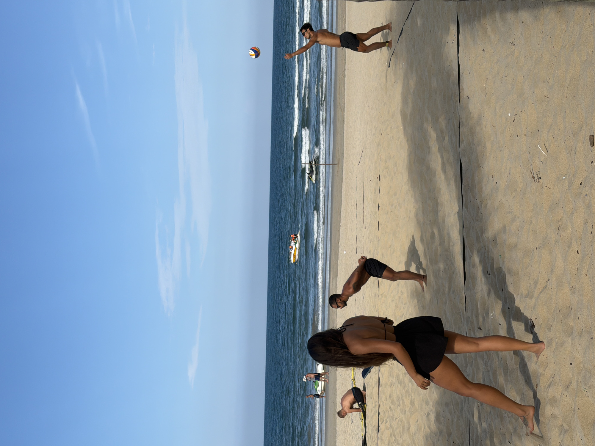 This image captures a vibrant beach scene in Đà Nẵng, Vietnam, on a bright, sunny day. The clear blue sky, dotted with a few wispy clouds, dominates the upper portion of the frame, meeting the expansive ocean. Gentle waves break along the shore, where several small boats carrying people are visible in the distance.
On the sandy beach in the foreground, people are engaged in a game of beach volleyball. Black boundary lines delineate the playing court on the light-colored sand. A shirtless man in black shorts is depicted mid-action, reaching out with an extended arm to connect with a colorful volleyball suspended in the air. Another shirtless man, also in black shorts, leans forward on the sand, suggesting he is in a ready or defensive position for the game. A woman with long dark hair, wearing a black top and black skirt or shorts, stands with her back partially to the camera, observing or participating in the activity. The bright sunlight casts distinct shadows on the sand, indicating mid-day conditions. The overall atmosphere is one of active recreation and enjoyment by the sea. No text is visible in the image.