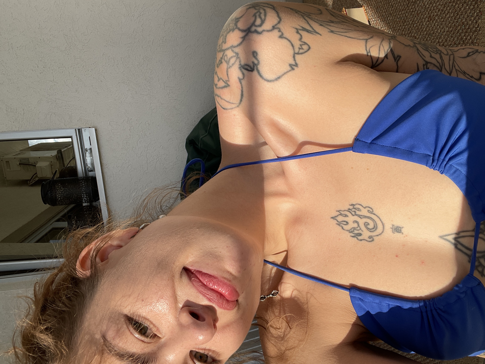 A low-angle selfie depicts a woman in a blue bikini, showcasing tattoos on her chest and upper body. The background reveals a section of a white wall and a mirror reflecting indoor furniture. The scene appears to be in Rafailovići, Montenegro, as suggested by elements in the background.