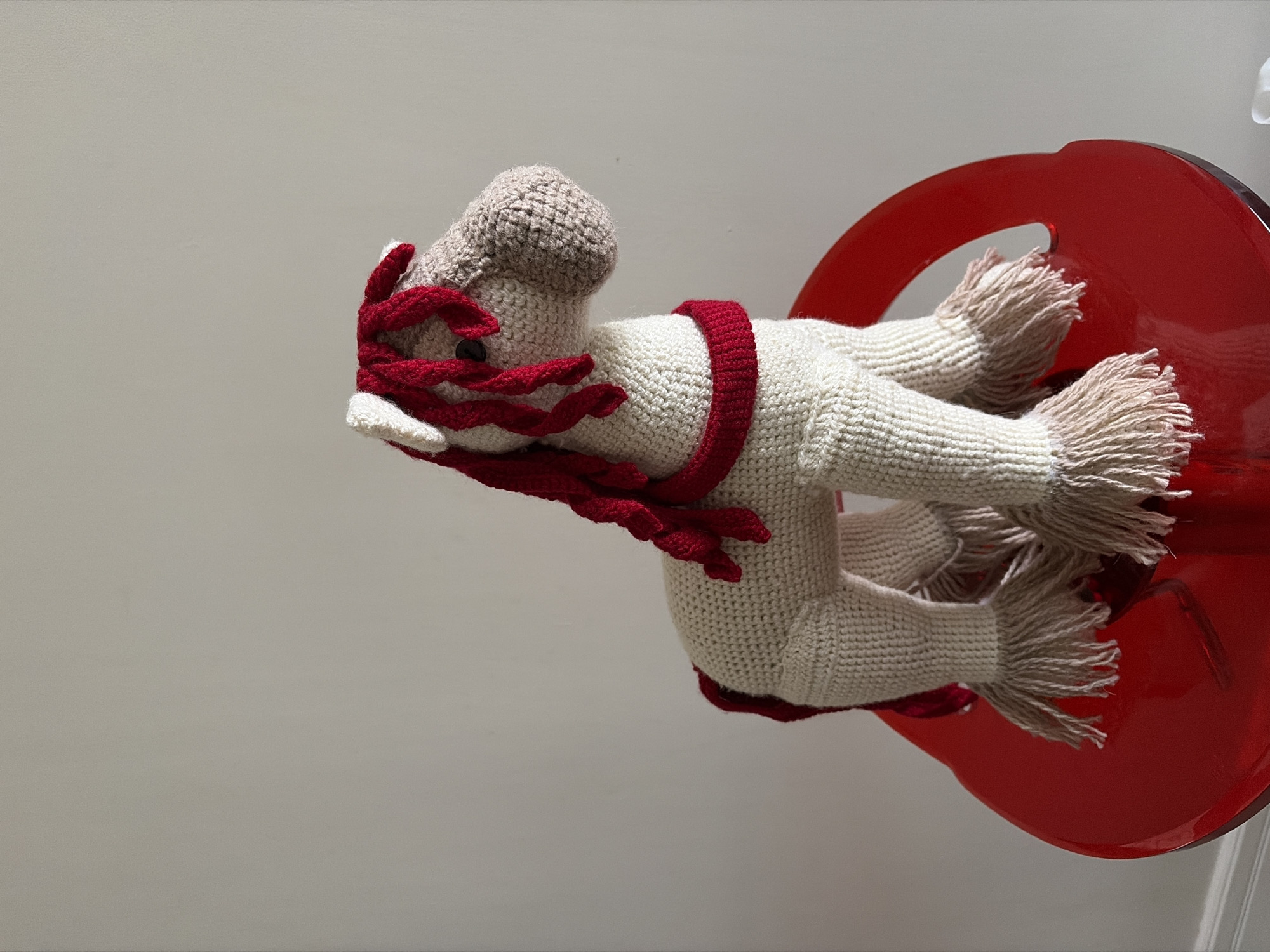 A crocheted horse toy, primarily cream-colored with a beige head and red mane and tail accents, is positioned on a bright red, molded plastic chair. The horse has a red band around its neck and fringed lower legs. The background is a plain, light-colored wall. The scene appears to be indoors. There is no visible indication of the city of Astana, Kazakhstan.