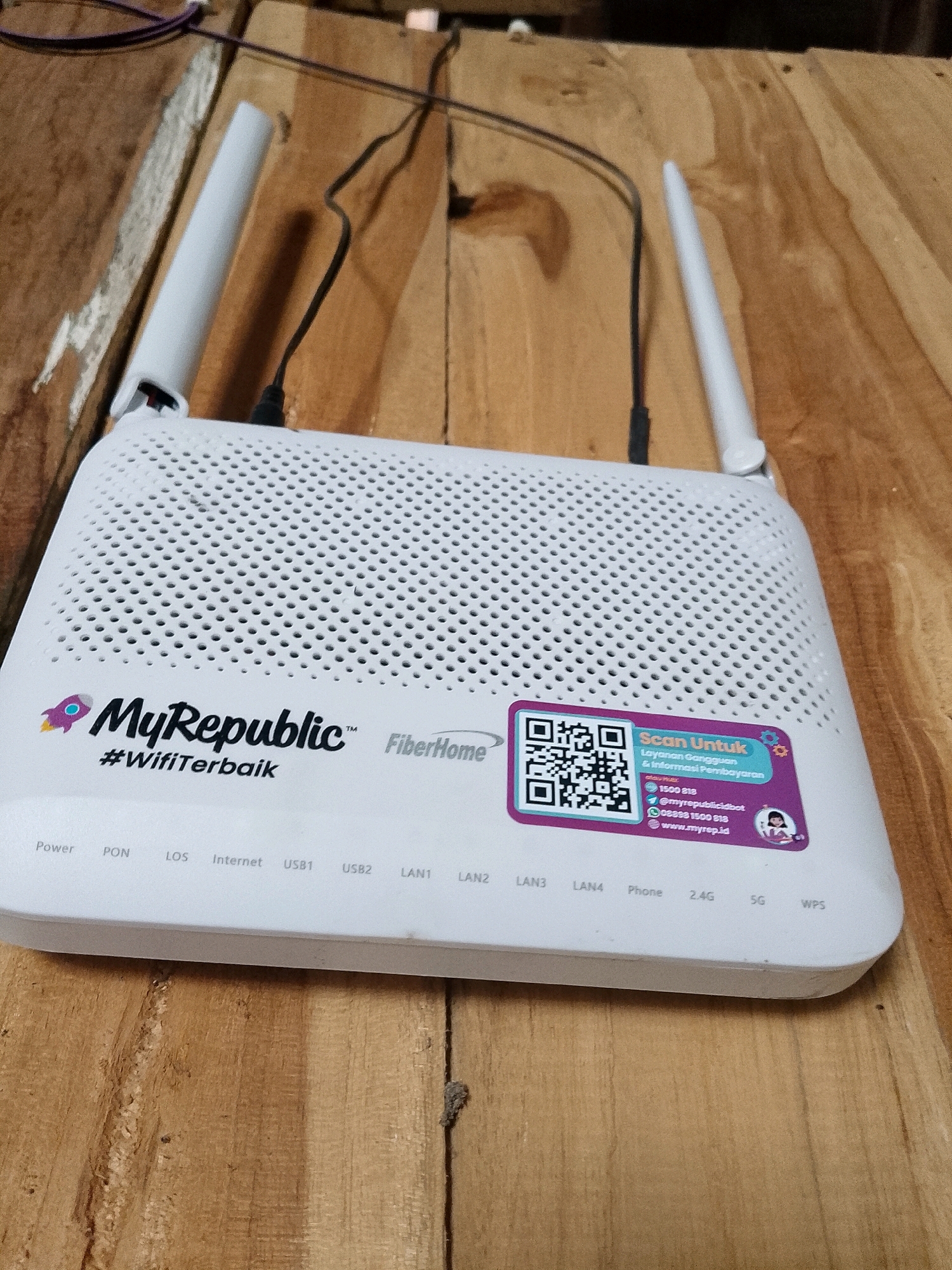 The image shows a white MyRepublic FiberHome router placed on a wooden surface, which appears to be an outdoor table made of planks. The router has two white antennas and several indicator lights labeled "Power," "PON," "LOS," "Internet," "USB1," "USB2," "LAN1," "LAN2," "LAN3," "LAN4," "Phone," "2.4G," "5G," and "WPS." A purple sticker with a QR code and contact information for MyRepublic is affixed to the right side of the router. The background is blurred, showing what seems to be more wooden planks and some dark cables. There are no people or animals visible in the image. The scene is lit by natural light. The location of Wadang, Indonesia, is not depicted or inferable from the image.