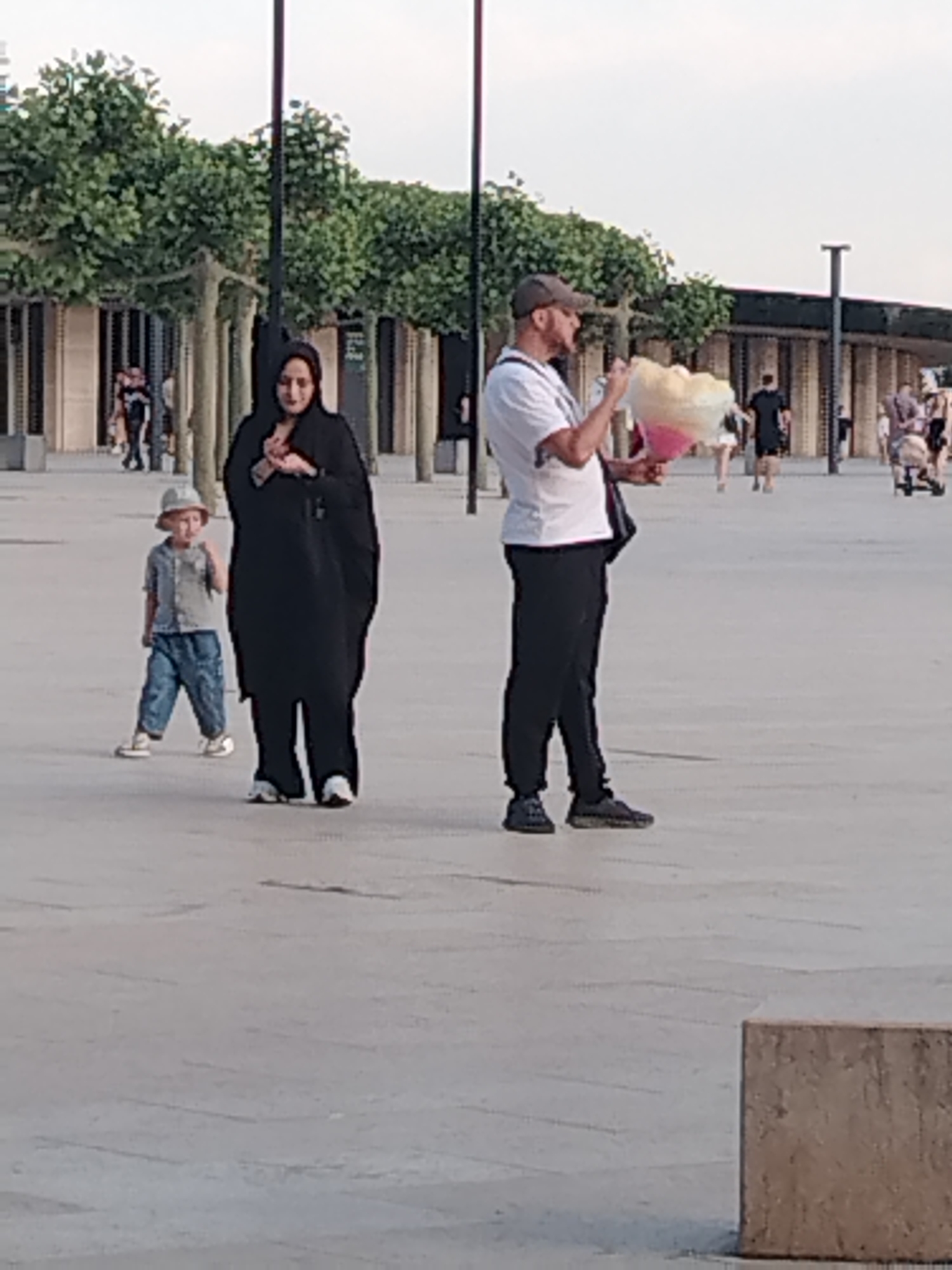 The image shows an outdoor scene with several people. In the foreground, a woman wearing a black abaya and a hijab is standing. To her left and slightly behind her, a young child in a light blue shirt, blue pants, and a light-colored hat is walking. To the right of the woman, a man wearing a white t-shirt, black pants, and a baseball cap is holding a large, pink and yellow cotton candy. He appears to be eating it. In the background, there are trees with green foliage, buildings with horizontal lines, streetlights, and other people walking. The ground is paved with large, light-colored tiles. The lighting suggests it is daytime, possibly late afternoon. The scene appears to be a public plaza or promenade. The provided information states the location is the city of Krasnodar, Russia, but this cannot be verified from the image itself.