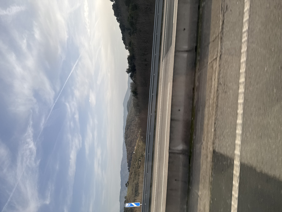 The image displays an elevated view of a highway or motorway in the region of Macedo de Cavaleiros, Portugal, under bright daylight. The foreground, shot from a moving vehicle, shows a dark asphalt road surface with a distinct dashed white lane marking. A low, grey concrete barrier, constructed from interlocking pre-cast sections, separates this foreground view from an elevated, multi-lane roadway. This elevated roadway features a lighter grey asphalt surface with multiple solid white lane markings. A silver metal guardrail runs along the left edge of this elevated road. Further left, partially visible beyond the guardrail, is a blue rectangular road sign with white pictograms, indicative of a service or amenity area (e.g., a fork and knife symbol). The background reveals a landscape of rolling hills and mountains, sparsely covered with brownish-green vegetation, suggesting a winter or early spring season. The sky is a light blue with scattered white clouds and a prominent, long white contrail from an aircraft extending diagonally across the upper left to the mid-right portion of the frame. Sunlight appears to be coming from the right, casting a shadow from the concrete barrier onto the lower foreground road. No individuals or other vehicles are distinctly visible in the immediate scene.