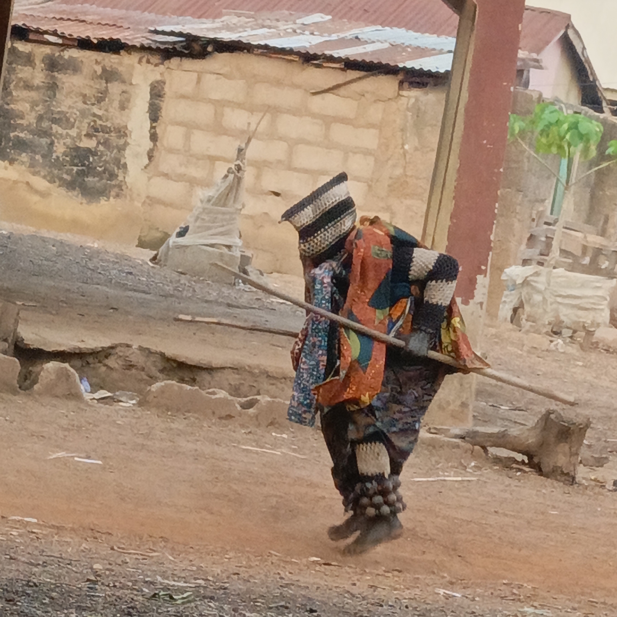 A figure wearing an elaborate, multicolored costume walks on a dirt road in Akwanga, Nigeria. The costume includes a striped, woven head covering, layers of patterned fabric, and beaded anklets or footwear. The figure carries a wooden stick horizontally across the shoulders. In the background are buildings made of earth-toned materials with corrugated metal roofs, suggesting a rural or underdeveloped urban area. The ground is a mixture of dirt and gravel.