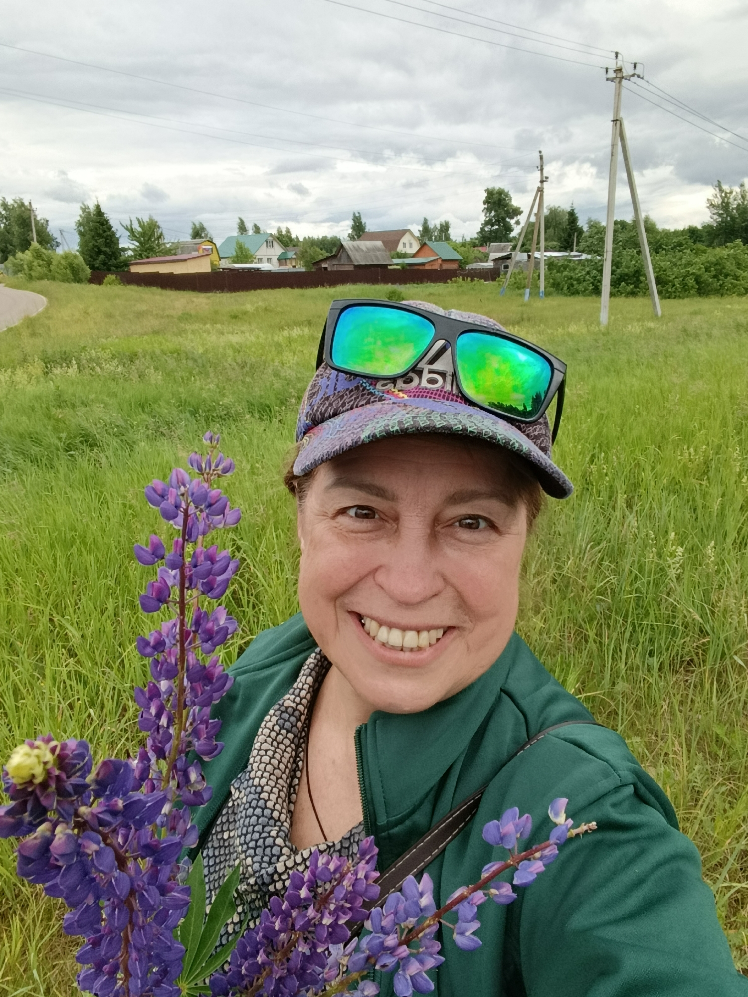A woman is taking a selfie in a field of tall green grass under a cloudy sky. She is wearing a patterned baseball cap with sunglasses perched on top, a green jacket, and a patterned shirt visible underneath. She is holding a bunch of purple lupine flowers in her left arm. In the background, there are houses with various roof colors, a dirt road on the left, and utility poles with wires. The scene appears to be in a rural or semi-rural setting, possibly in the village of Gribchikha, Russia, given the style of houses and the natural landscape.