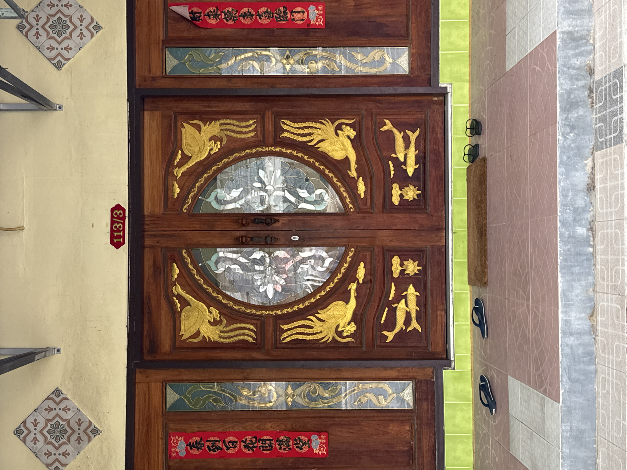 The media file shows a close-up view of a wooden door adorned with gold phoenix and fish motifs, typical of traditional Southeast Asian architecture. The door features ornate glass panels with intricate patterns, adding a decorative element. To the left of the door, a pale yellow wall is visible, decorated with a diamond-shaped tile and a vertical red sign with white text reading "113/3".
The setting appears to be the exterior of a building, possibly a residence or a shop, given the entrance context. The wall to the right of the door is tiled in shades of pink and grey, with a strip of green tiles visible between the wall and the door. Two pairs of black flip-flops are placed on the tiled floor near the entrance, suggesting a lived-in or functional space. The overall impression is one of a well-maintained entrance with decorative cultural elements. There is no visible indication of the time of day or weather conditions. The location context is Chalong, Thailand.