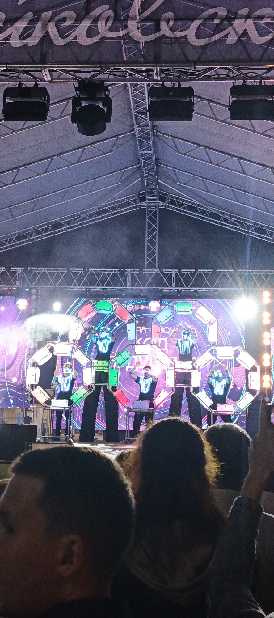 An outdoor music festival is taking place under a large tent. A stage is set up with a large LED screen in the background displaying a colorful, geometric pattern and text. Four performers are on stage, three of whom are on stilts, dressed in black outfits with illuminated geometric designs on their chests. They are interacting with large, octagonal structures that appear to be musical instruments, some of which are lit with green, white, and pink lights. The audience is visible in the foreground, with their backs to the camera, watching the performance. The overall atmosphere is that of a lively, nighttime event.