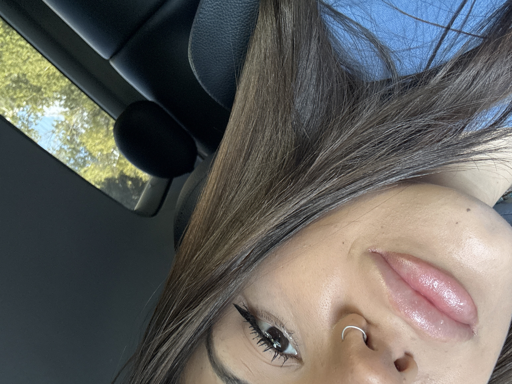 This is a close-up, angled selfie of a person, likely a woman, taken inside a car during daytime. The subject's head is tilted, creating a dynamic, almost upside-down composition, with long, dark brown hair cascading across the frame.
The visible part of the face includes one eye, prominent with a striking black winged eyeliner and voluminous dark eyelashes. The person has full, glossy pink lips, appearing moisturized or enhanced with a lip product, and a silver hoop septum piercing. The skin tone is warm, and the complexion appears smooth with a slight sheen, suggesting makeup application.
The setting is clearly a vehicle interior, characterized by dark-colored car seats or headrests and the car's ceiling structure. In the upper left portion of the image, a sunroof is visible, offering a glimpse of the outside. Through the sunroof, bright green foliage of trees and patches of blue sky indicate a clear, sunny day.
The activity captured is a self-portrait, presented with a casual and candid feel. There is no visible text in the image. While the provided context suggests Sofia, Bulgaria, the image itself does not contain any specific visual cues to confirm this geographical location, beyond the generic outdoor elements seen through the sunroof and the car interior.