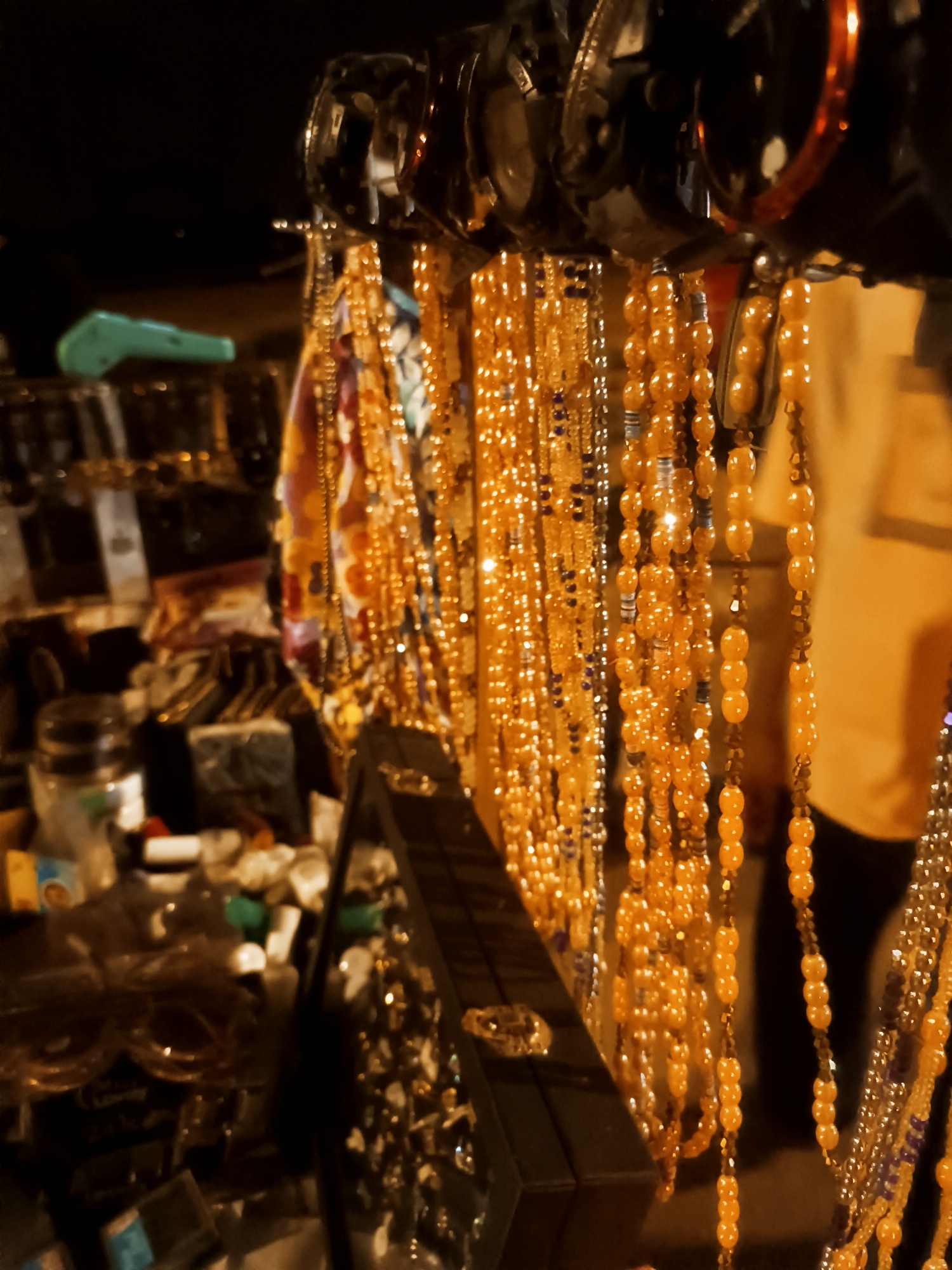 A photograph captures an outdoor marketplace scene in Bukuru, Nigeria. The focus is on a vendor stall showcasing an assortment of jewelry, mainly necklaces with orange beads hanging from racks. Sunglasses are displayed above the necklaces. Other items are scattered across the stall, adding to the marketplace atmosphere. A person in a yellow shirt is partially visible in the background. The overall lighting is dim, suggesting it is either dusk or night.