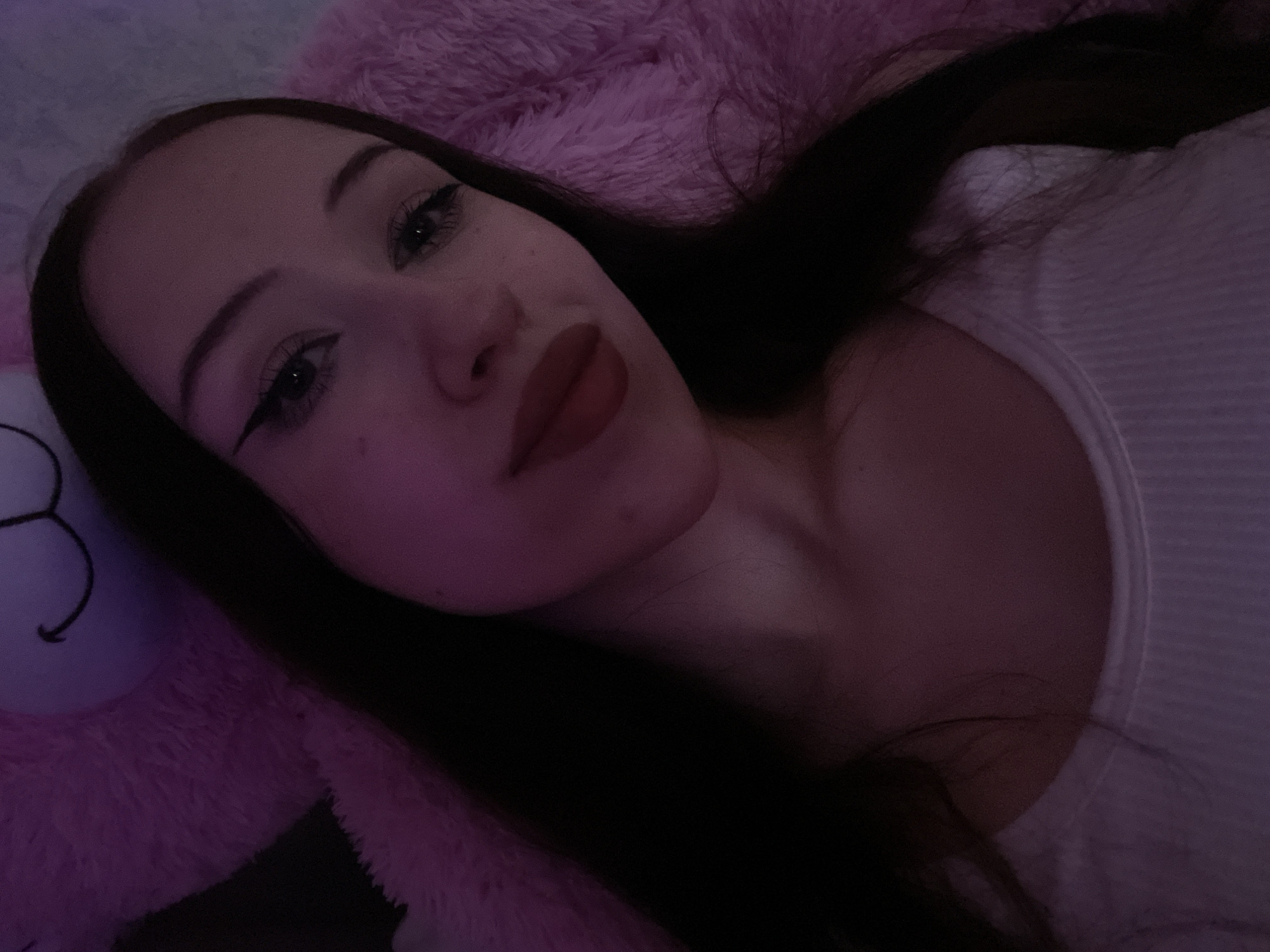 The image is a close-up selfie of a young woman lying down, possibly in bed, with a pink fluffy object, possibly a blanket or stuffed animal, behind her. She has dark hair and is wearing makeup, including winged eyeliner and red lipstick. The lighting is dim, with a pinkish hue, suggesting it might be nighttime or a dimly lit room. Her expression is neutral, and her eyes are looking towards the camera. The background is blurred, with only hints of the pink object and what appears to be a white top she is wearing. There is no visible text in the image.