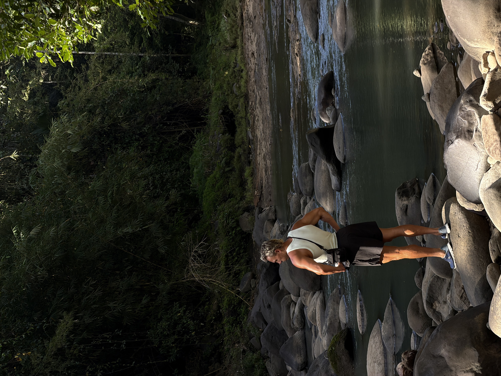 A person is standing on rocks beside a river. The river bank is covered with trees and vegetation. The location appears to be in Selemadeg Timur, Indonesia. The person is wearing a white tank top, black shorts, and blue shoes. They are standing with their hand on their hip. The river water is dark and flowing. The rocks are smooth and grey.