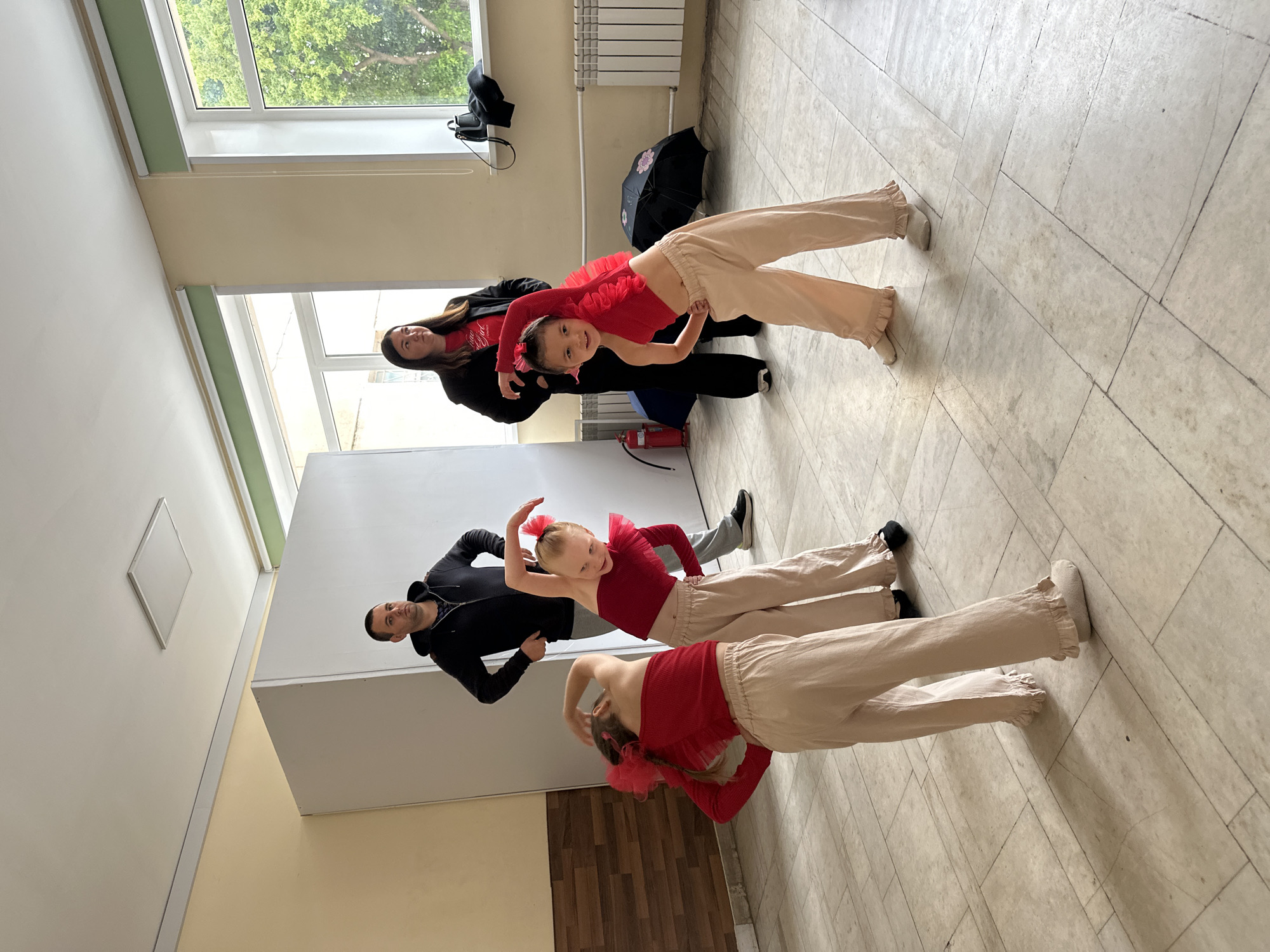 This image, taken in Chaikovsky, Russia, depicts a group of people inside a bright, well-lit room, likely a dance studio or rehearsal space. The photo is rotated 90 degrees counter-clockwise, showing the floor on the right and the ceiling on the left.
Three young girls, dressed in identical red ruffled tops, light beige baggy pants with ruffled ankles, and red hair bows, are the central subjects. They are performing synchronized dance poses, bending at the waist with one arm raised gracefully, all smiling towards the camera.
Four adults are also present. Two adult women wear black jackets over red shirts; one woman, partially visible, has "GRAND" text on her shirt, and another stands behind one of the dancing girls. Two adult men wear black hoodies; one observes near the children, and another is partially obscured behind a second dancing girl. The adults appear to be observing, supervising, or participating in the activity, with pleasant expressions.
The room features light-colored walls (white, beige, and a pale green border), large windows revealing lush green trees outside, and a tiled floor composed of light-colored squares and a section with a wood-like pattern. Visible objects include a white radiator under a window, a black umbrella, a black bag, and a red fire extinguisher. The natural light suggests it is daytime. The overall mood conveyed is one of joy and focused activity.