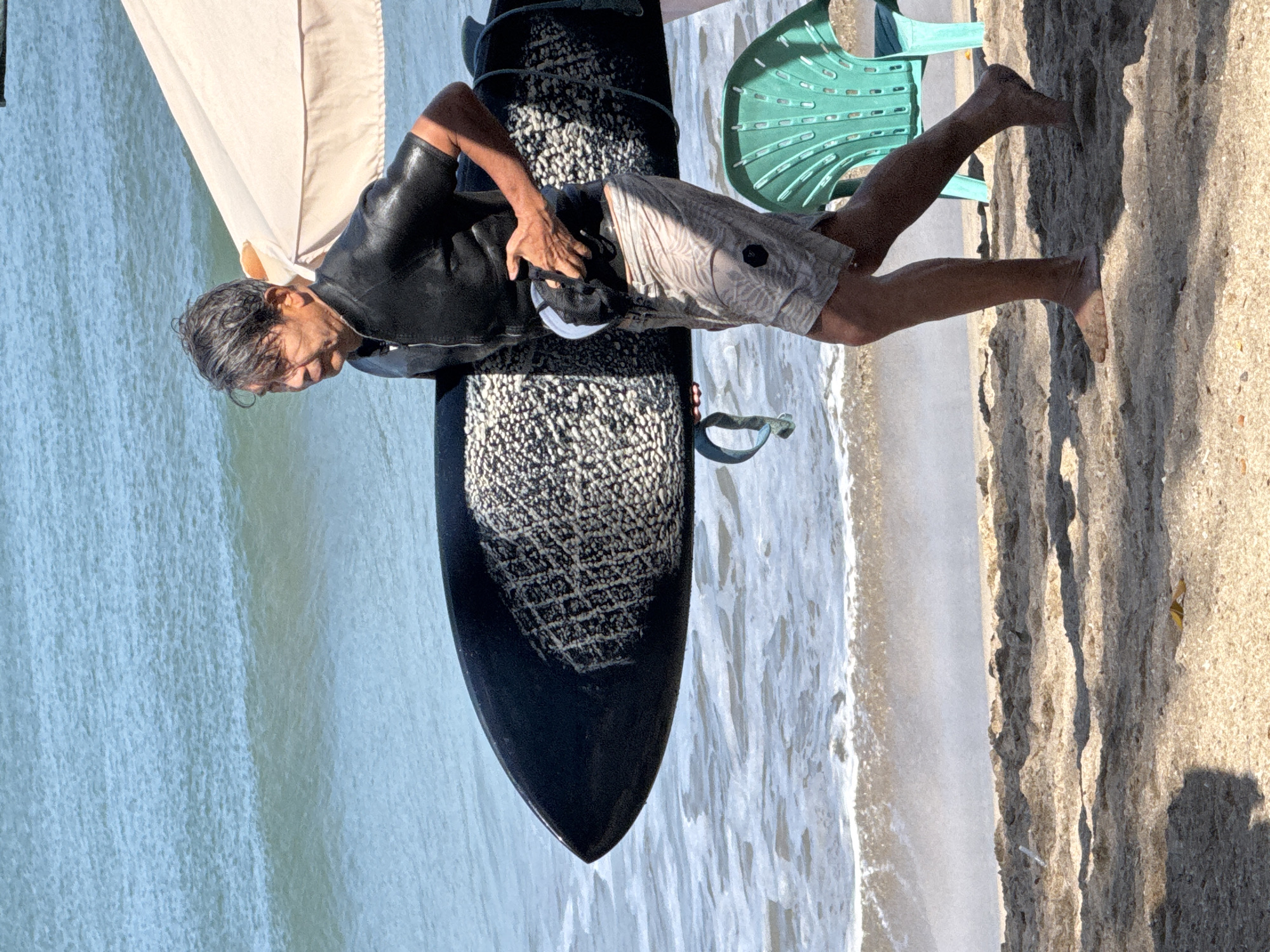 An older Asian man wearing a black short-sleeve wetsuit and patterned swim shorts walks along the sandy beach of Kuta, Indonesia, carrying a large black surfboard. The man is walking from left to right across the frame, with the surfboard held horizontally against his body. The ocean is visible behind him, with small waves breaking on the shore. A light-colored beach umbrella is positioned behind the man and to his right. In the foreground, on the right side of the image, a bright green plastic chair is partially visible. The sand is light brown and textured, with shadows cast by the umbrella and other unseen objects. The scene appears to be during the daytime, with bright sunlight.