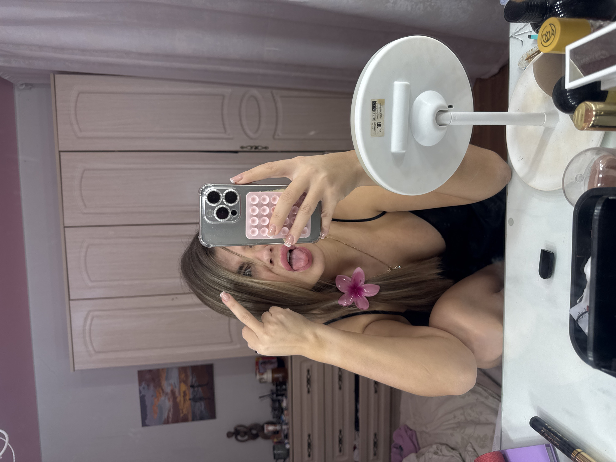 The scene depicts an indoor mirror selfie captured in a residential room, likely a bedroom or dressing area, in Chaikovsky, Russia. The primary subject is a female, seen from the waist up, with light brown hair, wearing a dark top and a necklace with a prominent pink flower charm. She is looking directly towards the camera, holding a smartphone with a clear case and a pink pop-socket style grip in her right hand, while simultaneously extending her left middle finger upwards and protruding her tongue from an open mouth.
In the immediate foreground, a white table surface is visible, upon which a white, circular standing makeup mirror is positioned. The table is cluttered with various objects, including what appears to be a black marker, a black marker cap, and multiple cosmetic bottles or containers.
The background reveals a furnished room. To the left of the subject, a light-colored wooden dresser with multiple drawers is visible, adorned with various small items and a dark wooden sculpture. Above the dresser, a framed artwork depicting a sunset over a body of water with two silhouetted figures hangs on a light-colored wall. A rumpled bed with light-colored bedding is partially visible in the lower left corner. Centered in the background is a large, light-colored wooden wardrobe featuring two doors, each with an arched panel design. To the right of the wardrobe, a light-colored curtain or drapery is present. The room's wall color appears to be a soft pink or lavender hue.