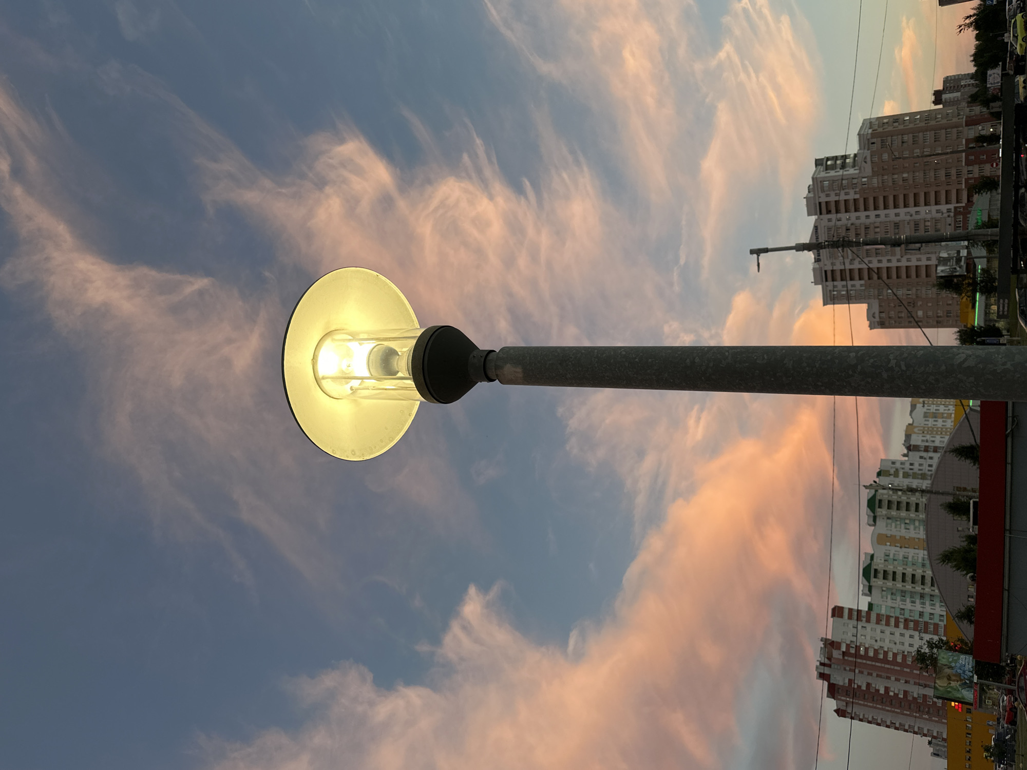 A low-angle, vertical medium shot captures a solitary streetlamp against a dramatic sky painted with wispy, pink-tinged clouds during what appears to be either dawn or dusk. The lamp itself, illuminated by a bright, warm bulb, is centered in the frame, its pole extending downwards out of view. In the background, a cityscape unfolds with multi-story apartment buildings characterized by varied architectural styles and colors, some featuring prominent balconies and windows. Power lines crisscross the scene, connecting the buildings. Below the buildings, hints of a street or parking area are visible, with a few cars and what might be commercial signage. The overall impression is of an urban setting at a transitional time of day, with the illuminated streetlamp standing as a solitary sentinel against the vibrant sky.