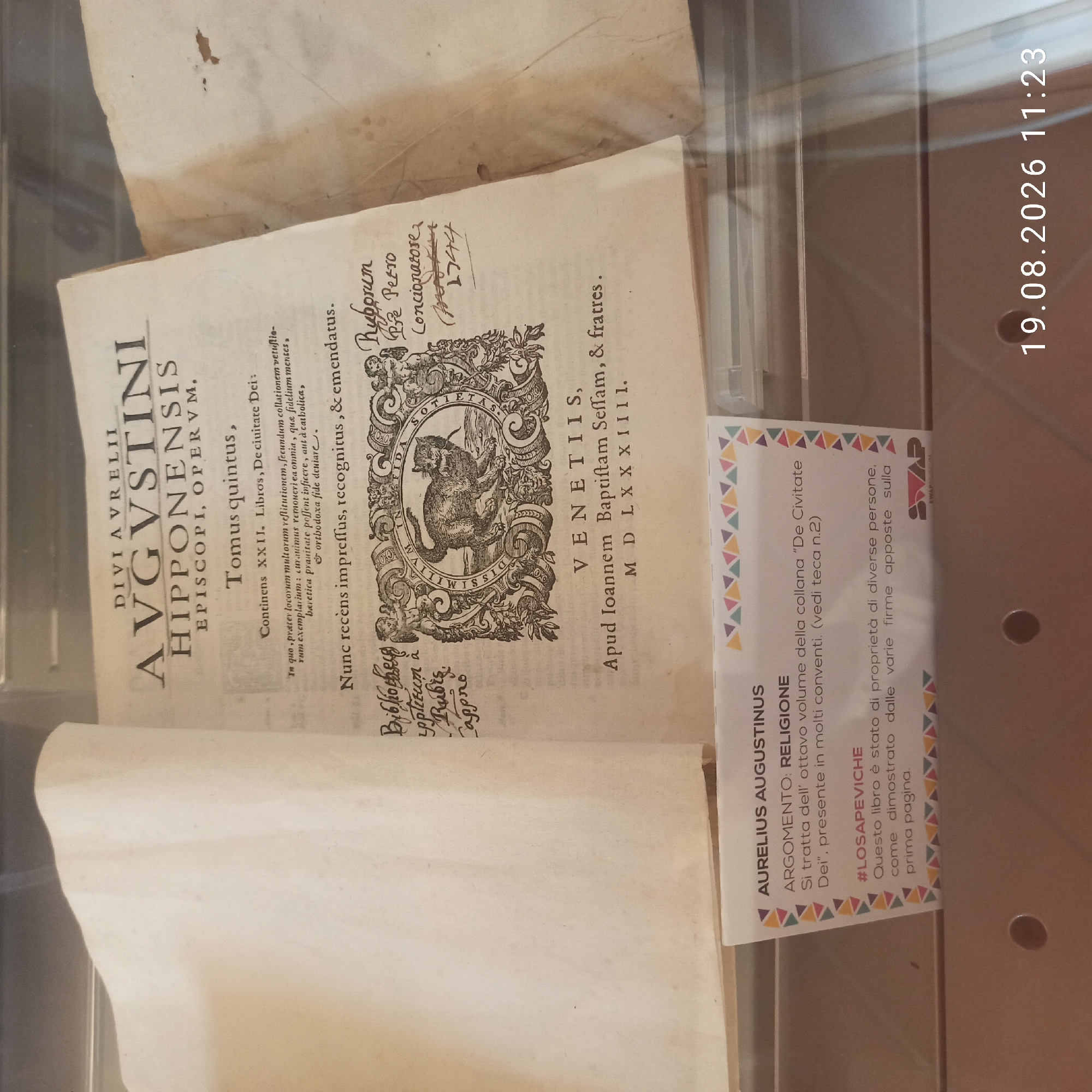The image displays an open, old book on a surface. The book is primarily composed of yellowed pages with printed text and an intricate illustration in the center. The illustration features a cat within an ornate, circular frame. Above the illustration, the book's title is prominently displayed: "DIVI AVRELII AVGVSTINI HIPPONENSIS EPISCOPI, OPERVM." Below the title, "Tomus quintus," indicates it's the fifth volume. Further text includes Latin phrases and what appears to be handwritten notations on the right side. Below the illustration, the location of publication is given as "VENETIIS, Apud Ioannem Baptiftam Seffam, & fratres. M D L X X X I I I." This indicates the book was published in Venice in 1583.
On the left side of the book, there are handwritten notes that appear to be library or ownership marks: "Bibliotheca," "oppoſitum à," "Rubis," and "lappino."
A small informational card is placed in front of the book. The card has a colorful triangular border and text in Italian. It reads: "AURELIUS AUGUSTINUS ARGOMENTO: RELIGIONE Si tratta dell' ottavo volume della collana 'De Civitate Dei', presente in molti conventi. (vedi teca n.2) #LOSAPEVICHE Questo libro è stato di proprietà di diverse persone, come dimostrato dalle varie firme apposte sulla prima pagina." This translates to: "AURELIUS AUGUSTINUS SUBJECT: RELIGION This is the eighth volume of the collection 'City of God', present in many convents. (see case n.2) #WELOSAKENOW This book belonged to several people, as shown by the various signatures on the first page."
The background appears to be a display case or shelf. In the bottom right corner of the image, a date and time stamp is visible: "19.08.2026 11:23".
The image does not provide any information about the city of Ruvo di Puglia, Italy, nor does it contain any people or specific actions. The scene is static and focused on the book and its accompanying information card within a display environment.