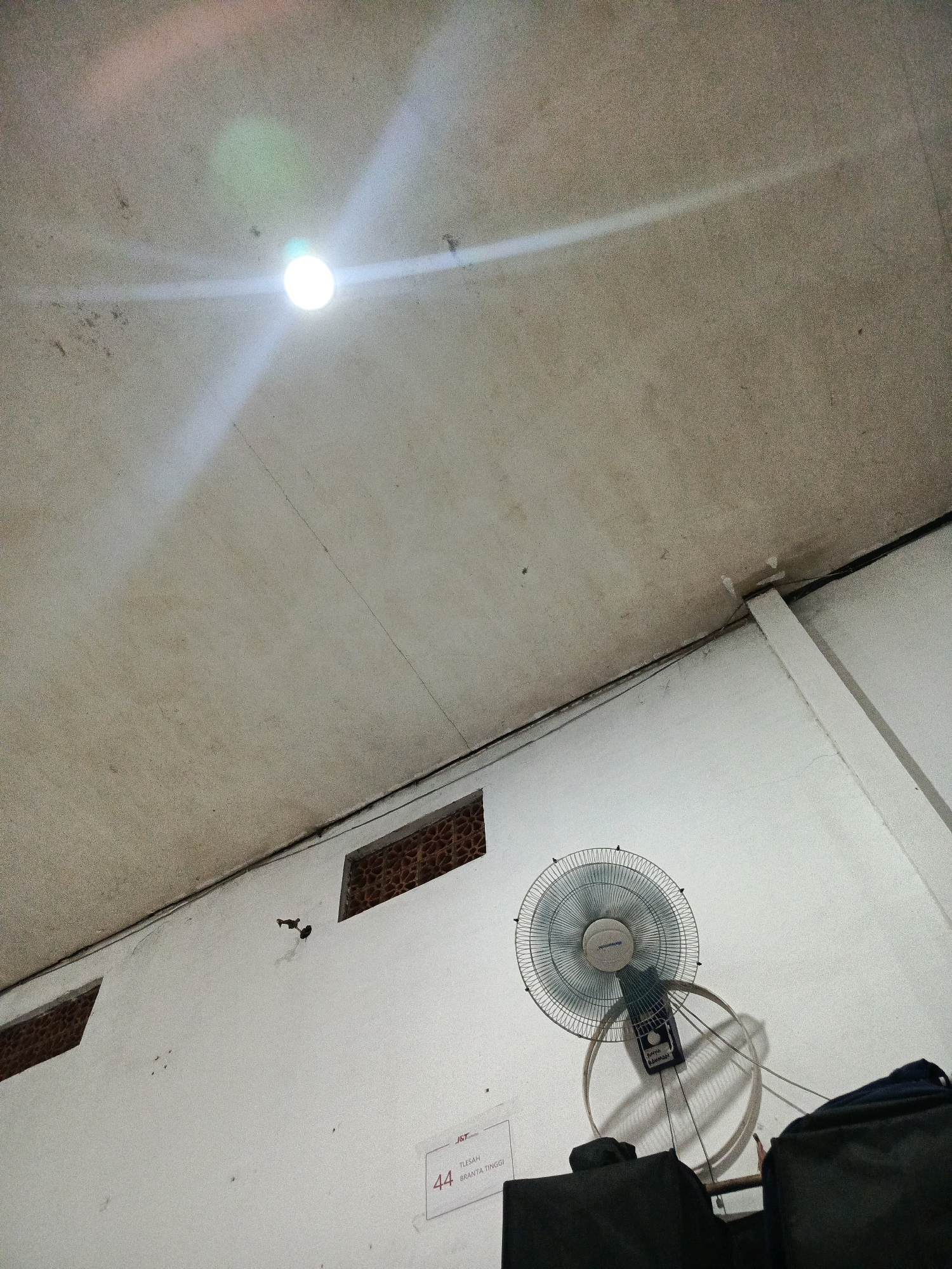 The image shows a ceiling with a bright light fixture emitting glare. Below, a white wall is visible with two small, rectangular openings with red brick grating. A fan is mounted on the wall, casting a shadow. In the lower right of the image, part of a blue backpack and a black bag are visible, suggesting a storage or transportation context. To the left of the fan, a sign with the number "44" and Indonesian text "TLESAH BRANTA TINGGI" is affixed to the wall. This suggests the location is likely within a building in Pamekasan, Indonesia, possibly a warehouse, storage facility, or transportation hub, given the signage and bags. The overall lighting is dim, with the ceiling light being the primary source.