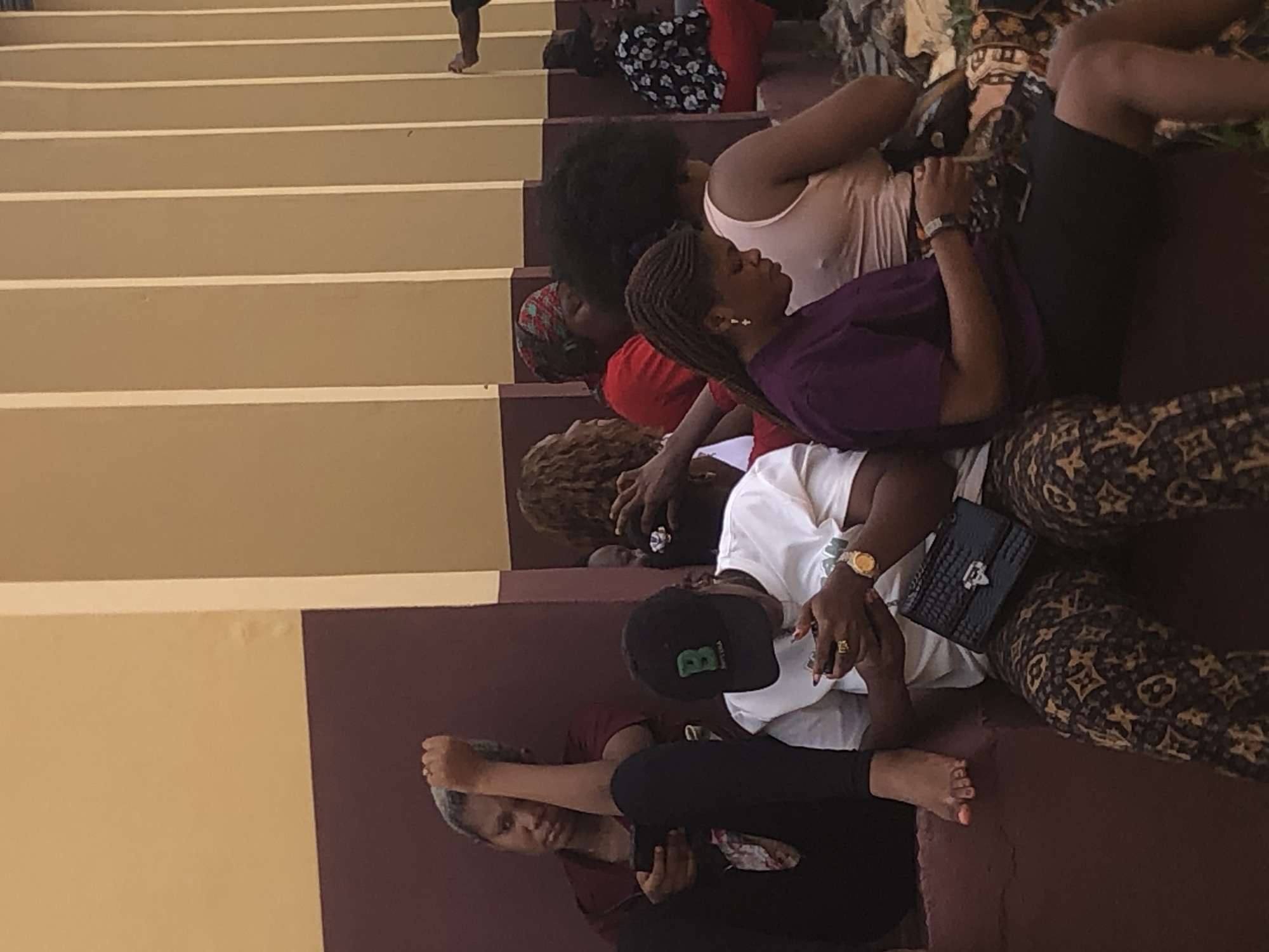 People are sitting on the stairs of a building in Nsugbe, Nigeria. They are various ages and are wearing casual clothing. Some are interacting with their phones. The scene is inside, evidenced by the walls and ceiling.