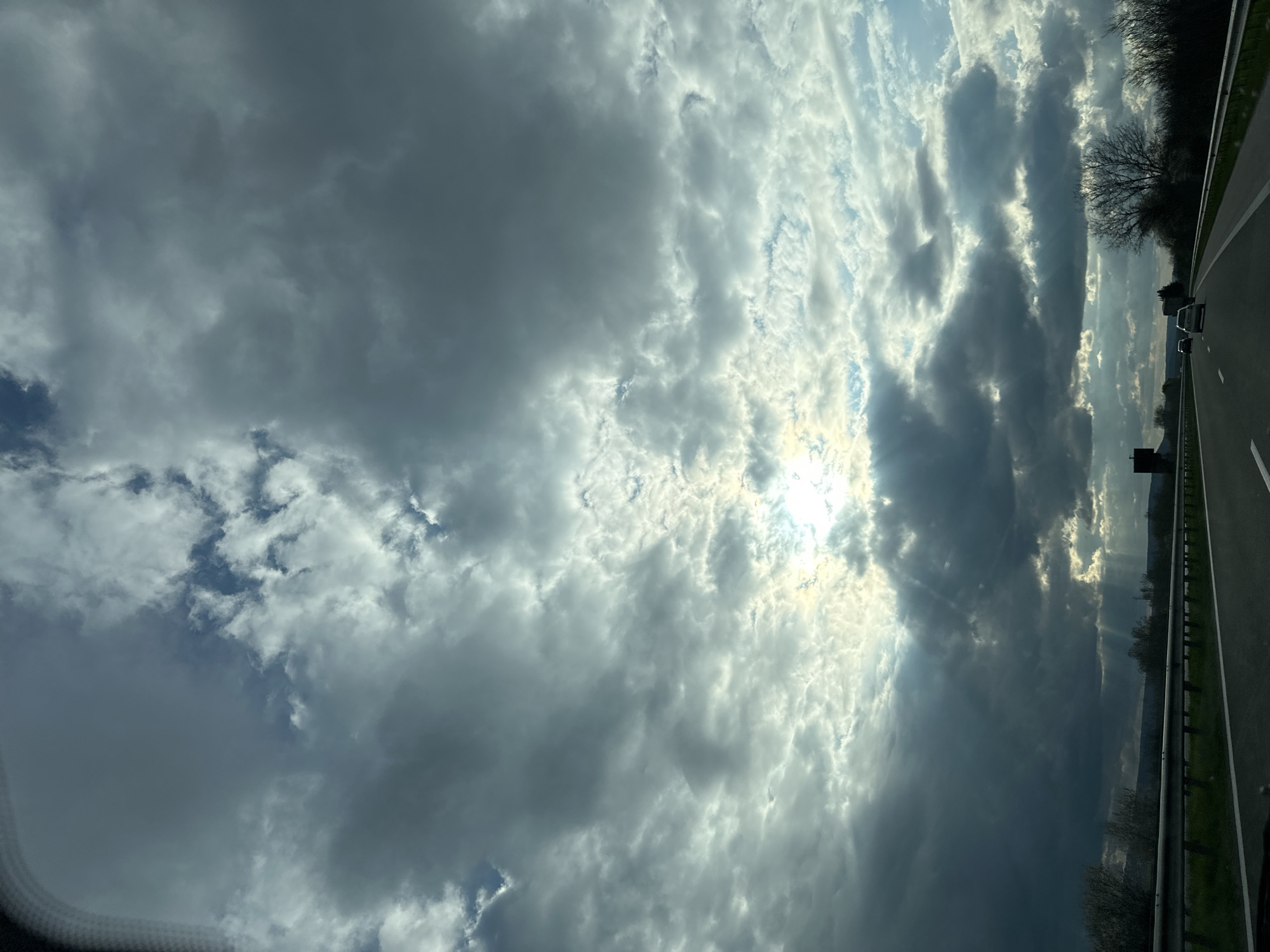 The image shows a view from a vehicle moving on a highway. The sky is filled with clouds, ranging from light and wispy to dark and dense. The sun is visible through a break in the clouds, creating rays of light. To the right of the frame is the highway with a safety barrier, trees, and some other vehicles in the distance. Given the request for location context, it is implied that the image was taken near Gabarnitsa, Bulgaria. However, this context does not change the visual description of the provided image.