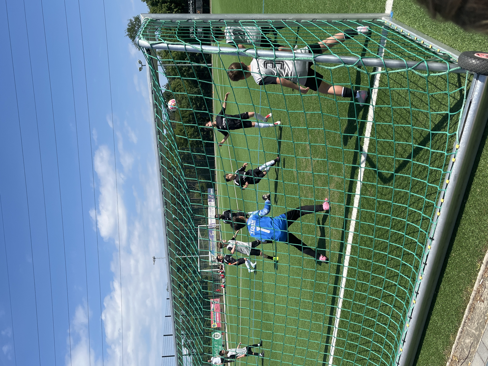 A soccer game is in progress on a green field in Kelsterbach, Germany. Players in black and white uniforms and a goalie in a blue uniform are on the field. One player in a black uniform is on the ground, and a soccer ball is in the air. The view is from behind a green soccer net with a bright blue sky above.