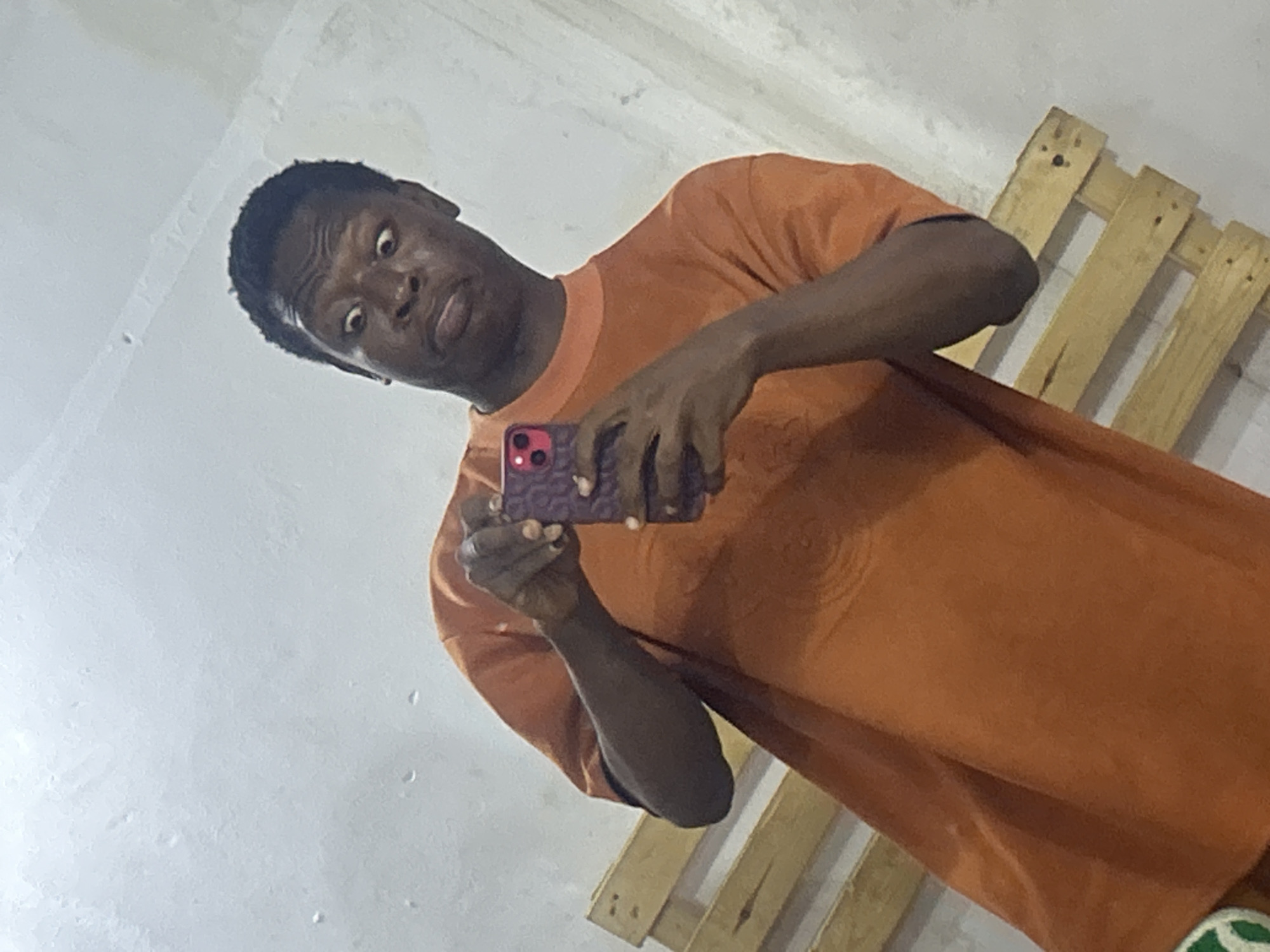 A selfie angled upward shows a person with dark skin and short, black hair, wearing an orange t-shirt. The person is holding a smartphone with a pink case, their face showing a somewhat surprised or quizzical expression. The background consists of a white wall and a wooden structure made of planks. The city and specific location are unknown.
