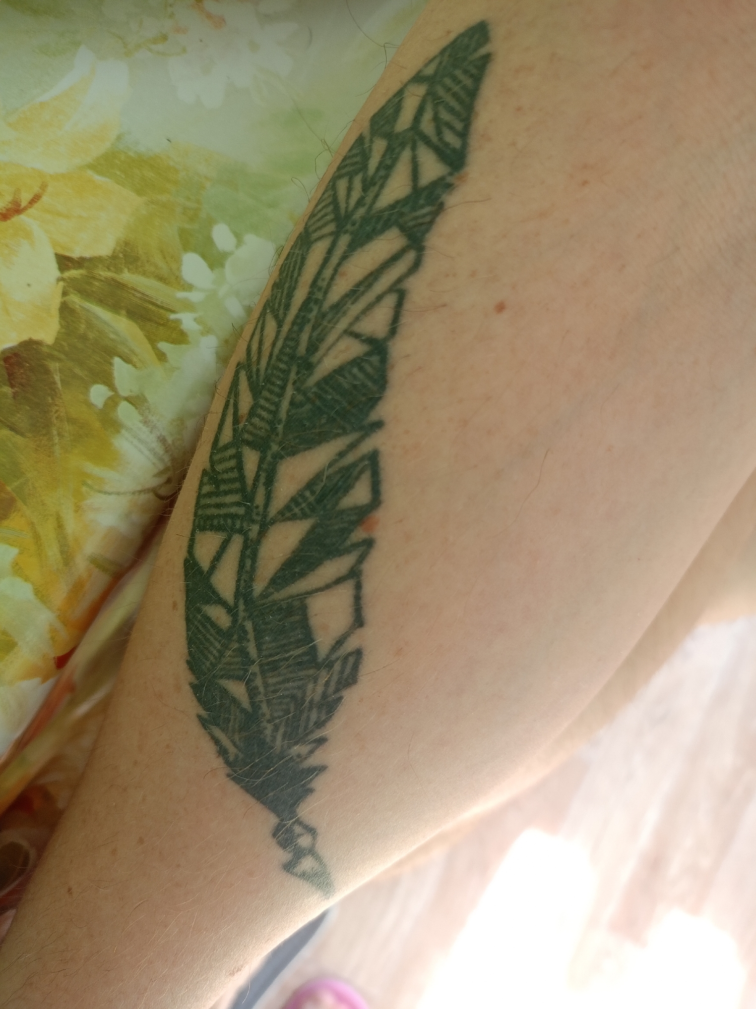 An arm with a dark green feather tattoo is positioned diagonally across the frame, resting on a patterned fabric with floral motifs. The tattoo features geometric designs within the feather. The skin exhibits fine hairs and minor discolorations. The background consists of the aforementioned fabric and a bright, out-of-focus area suggesting an indoor environment with lighting. No location in Moscow, Russia, is identifiable from the image.