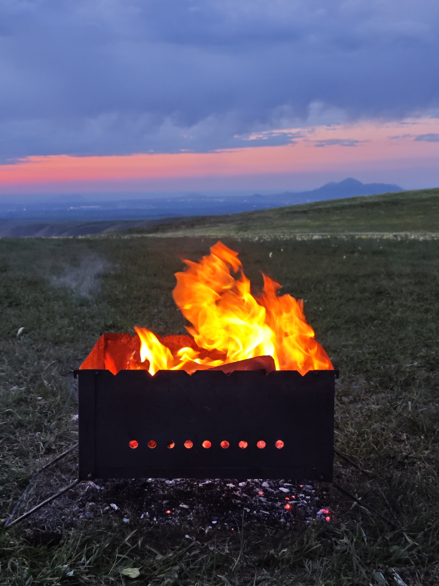 A metal brazier filled with burning wood stands on a grassy field. The fire produces bright yellow and orange flames. Below the brazier are embers and ash. In the background, a landscape with rolling hills and a distant mountain range is visible under a sky transitioning from dark blue-gray at the top to a band of pink and orange on the horizon. The location is near the city of Lermontov, Russia.